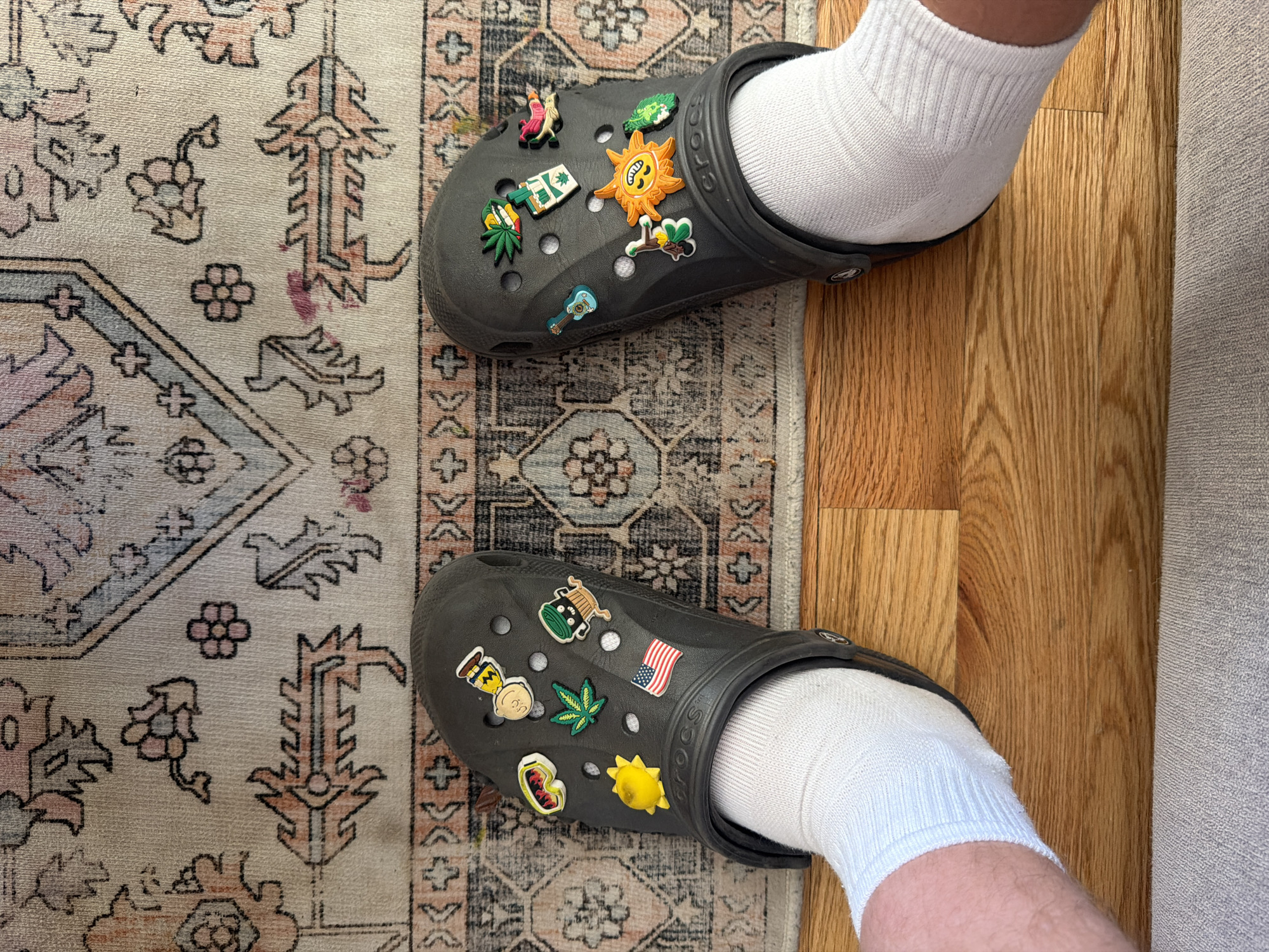 The image depicts a person wearing gray Crocs with white socks standing on a patterned rug and wooden flooring, in an indoor setting. The Crocs are adorned with various decorative charms. One Croc has a charm resembling a sun and the other has a charm resembling the American flag. The flooring appears to be hardwood. The location in Morton Grove, United States of America is not determinable from the image alone.
