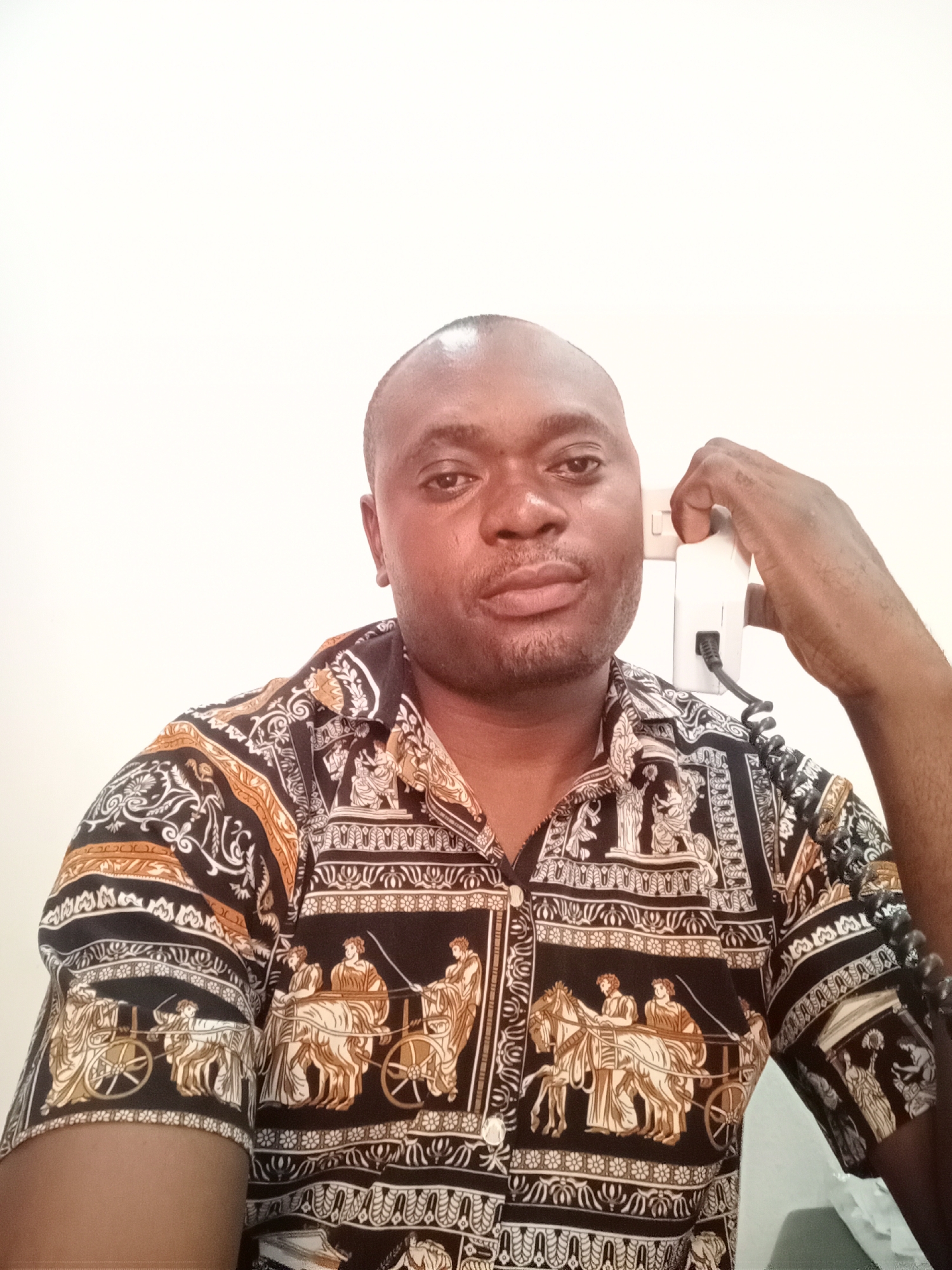 A man with dark skin, a shaved head, and a neutral expression is captured in a chest-up portrait, looking directly at the camera. He is indoors, against a plain white wall, likely in Gwarinpa, Nigeria.
He is wearing a short-sleeved button-up shirt featuring an elaborate pattern primarily in black, gold-brown, and white. The intricate design on the shirt depicts scenes reminiscent of ancient Greek or Roman life, with chariots pulled by horses and figures dressed in robes, interspersed with decorative borders and scrollwork.
With his right hand, he holds a white telephone receiver to the side of his head, possibly as a prop for the photograph or pretending to be on a call. A black coiled cord is visible extending from the receiver. His left arm is also visible, showcasing the pattern on the sleeve. The overall impression is a posed photograph.