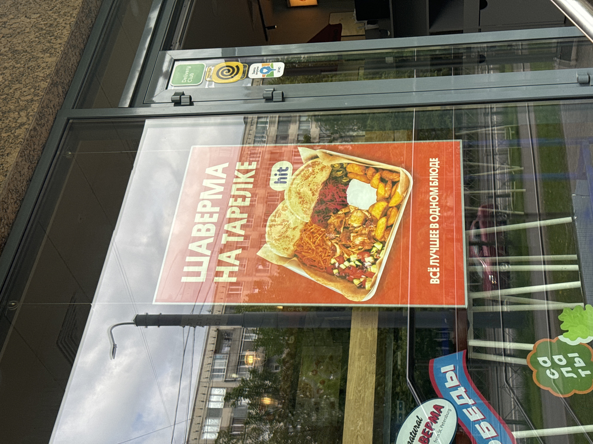 An image, rotated on its side, captures the storefront window of a food establishment in Saint Petersburg, Russia, during daytime. The window serves as a display for several advertisements and informational stickers.
The most prominent subject is a large advertisement for food, depicting "ШАВЕРМА НА ТАРЕЛКЕ" (Shawarma on a plate) with a "hit" label, and text that reads "ВСЁ ЛУЧШЕЕ В ОДНОМ БЛЮДЕ" (All the best in one dish). The food platter includes flatbread, meat, fried potatoes, and various salads. Below this, another sticker, partially visible, advertises "natural ВЕРМА from St. Petersburg" (likely referring to Shawarma), "ОБЕДЫ" (Lunches), and "са ла ты" (Salads).
Multiple service stickers are affixed to the top of the window frame, including "Delivery Club," "Здесь вкусно" (Here is tasty), and "2ГИС" (2GIS, a mapping service).
The window's reflective surface shows a clear blue sky, a tall lamppost, and the facades of buildings, suggesting an urban street setting. The sunlight indicates it is daytime with good weather. No people are visible, and the scene focuses entirely on the commercial signage and its reflections. The overall impression is that of a casual eatery promoting its offerings and delivery options.