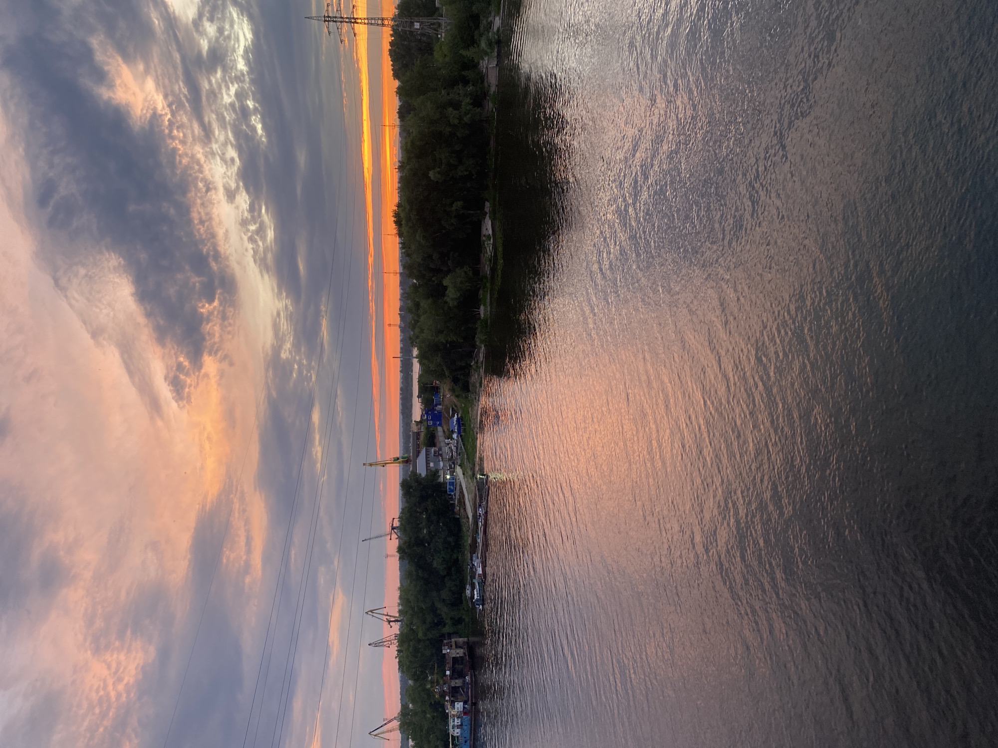 This is a photograph taken from a bridge over the Volga River in Samara, Russia, during sunset. The sky is filled with dramatic clouds tinged with orange and pink hues from the setting sun. In the distance, along the riverbank, are industrial cranes, buildings, and dense green trees. Several barges and smaller boats are docked in the river. The water reflects the colors of the sky, creating a shimmering surface. Power lines and pylons are visible stretching across the frame, indicating the presence of urban infrastructure. The scene captures a blend of industrial activity and the natural beauty of a sunset over the river.