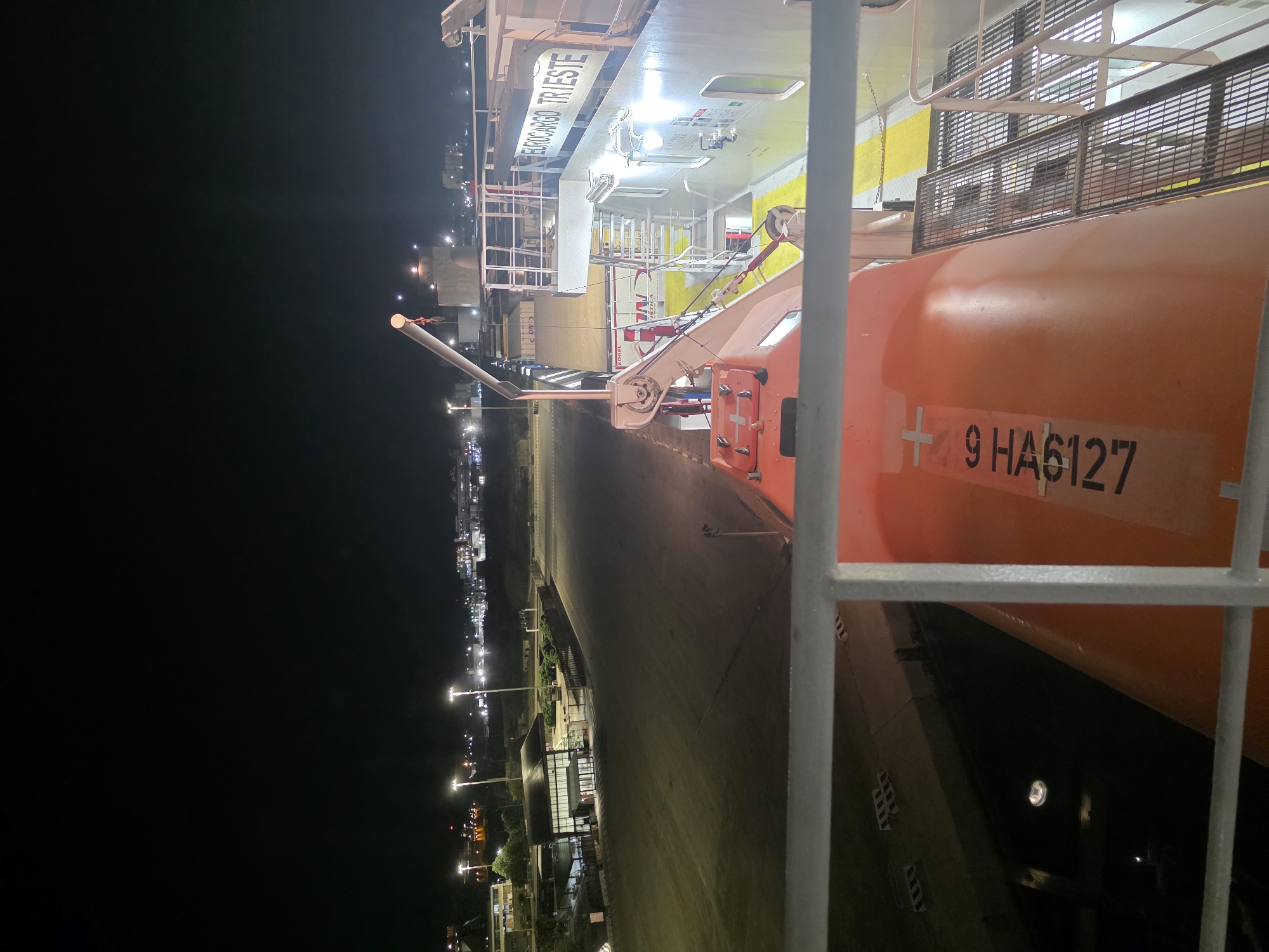 The image is a night time photograph taken from the deck of a ship docked in Brindisi, Italy. The foreground shows a metal railing and the orange hull of a life raft with the registration number "9 HA6127" printed on its side. Behind the life raft, a section of the ship's superstructure is visible, including a crane and the name "EUROCARGO TRIESTE" emblazoned on the side. In the middle ground, a paved area, likely a dock or ferry terminal, is illuminated by bright streetlights. Several buildings and structures are visible in the background, as well as distant city lights, indicating an urban or port area. The overall impression is of a commercial port at night.