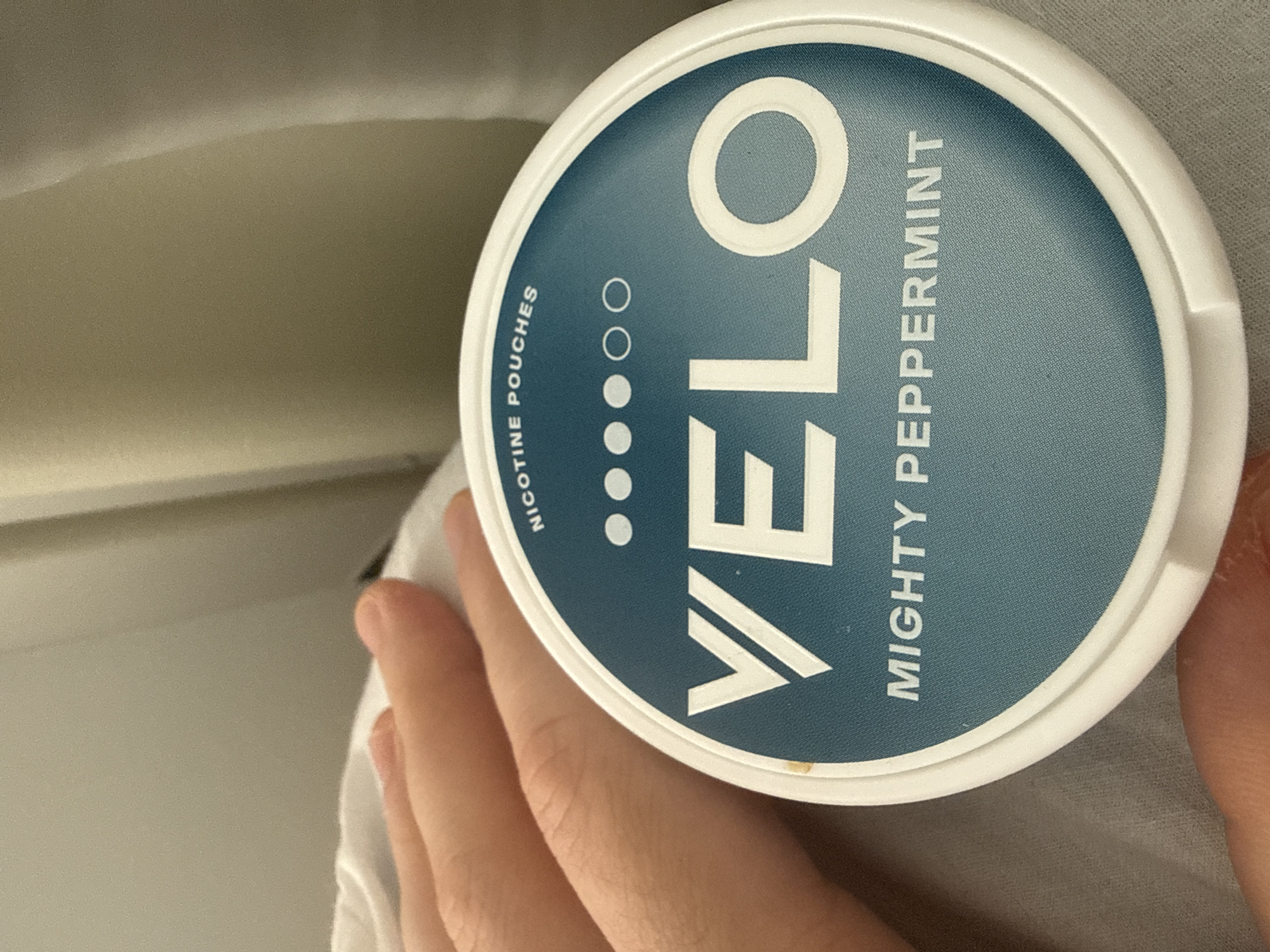 A close-up shot depicts a white, circular container with a blue label for "Velo Mighty Peppermint" nicotine pouches. The container is held in a person's hand. The background is blurred, showing a light-colored surface and a glimpse of a white material, possibly fabric. There is no indication of the location in Vynnyky, Ukraine, or any other city.