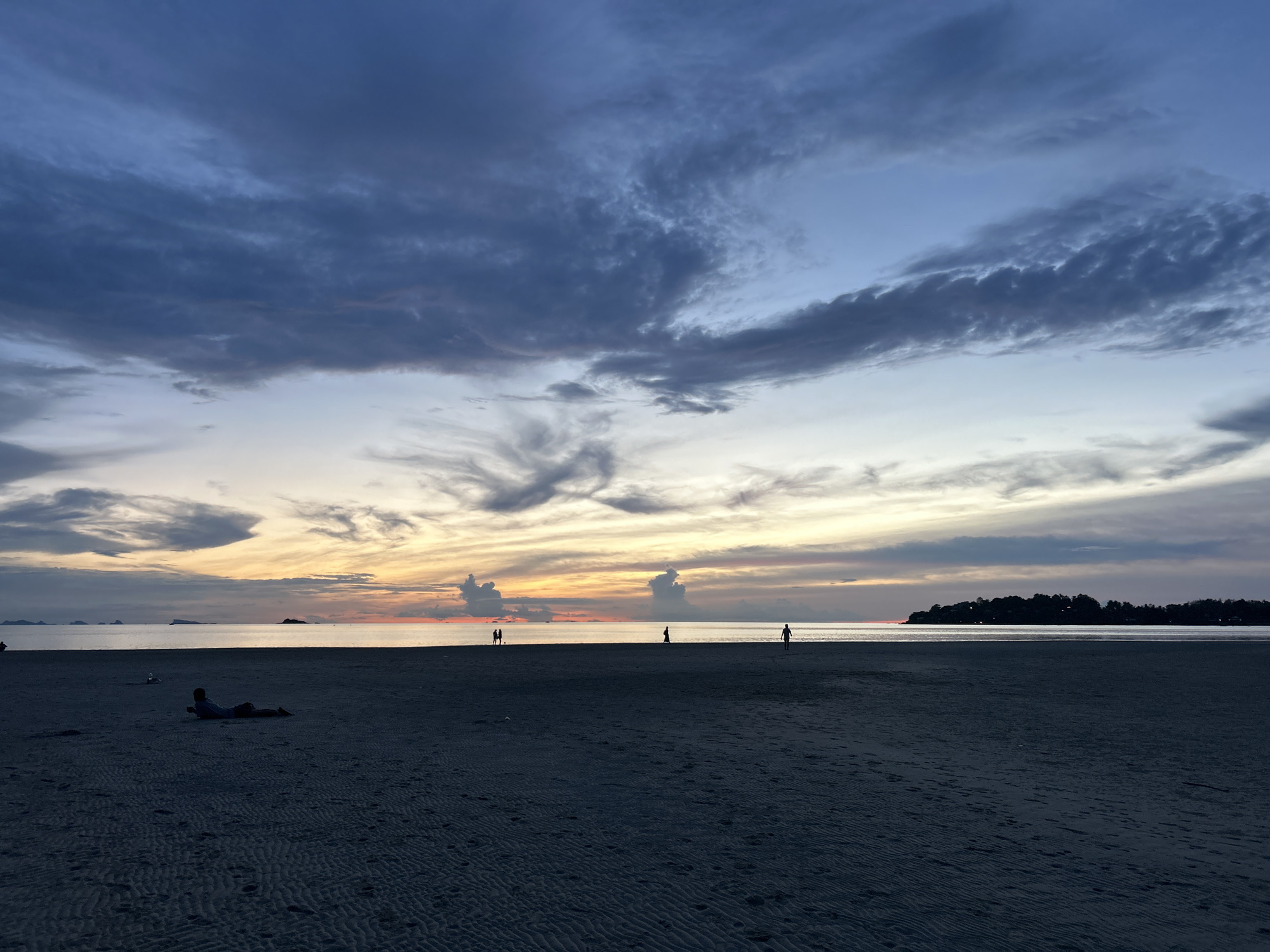 This image depicts a beach scene at dusk on Koh Phangan, Thailand. The sky is filled with dramatic clouds, transitioning from deep blues at the top to a warm orange and yellow glow near the horizon where the sun has set. The ocean is calm and stretches out to meet a distant, dark silhouette of landmasses, likely other islands or parts of Koh Phangan. The foreground is a wide expanse of dark, textured sand, showing ripples from the receding tide and scattered footprints.
On the beach, several figures are visible. In the left mid-ground, a person is lying on their stomach, facing away from the camera, relaxed on the sand. Further to the right, in the mid-ground, two figures stand close together near the water's edge, appearing to be conversing or interacting. Another solitary figure walks further out on the beach, and another is seen in the far right mid-ground. The overall atmosphere is serene and tranquil, capturing the end of a day with soft, diffused light.