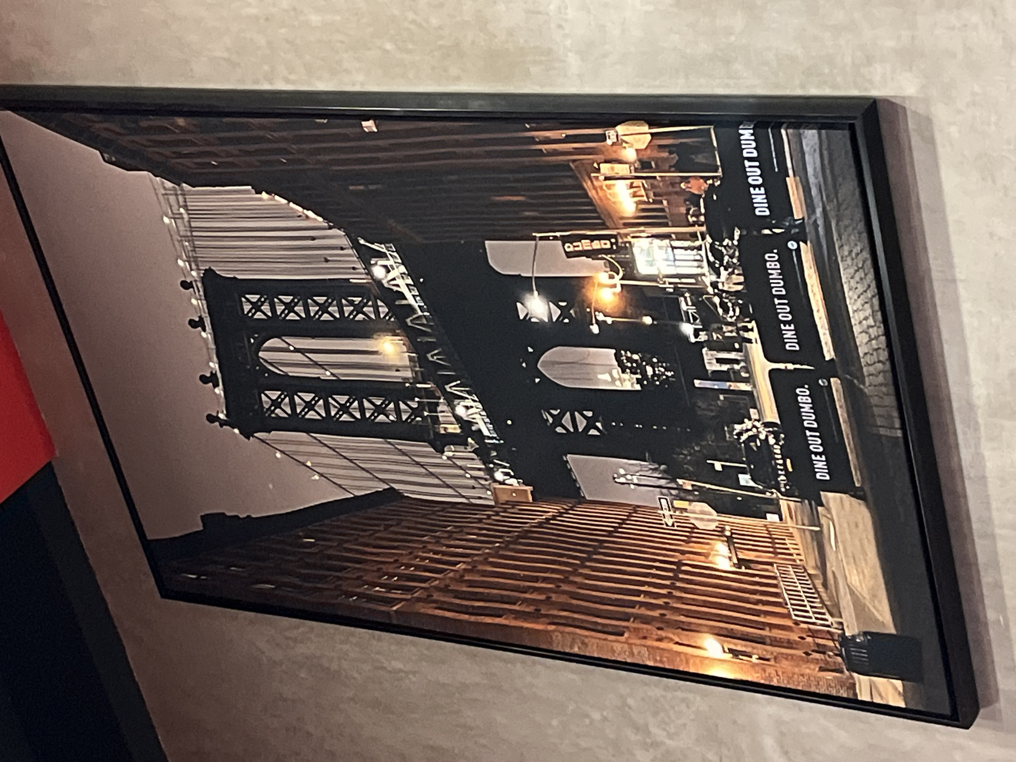 Here is a detailed description of the image:
The image shows a framed print hanging on a light-colored wall. The print depicts a nighttime urban scene with a prominent suspension bridge. The bridge has a latticework of steel beams and cables. "DINE OUT DUMBO" is written on the right side of the image. Below the bridge structure, there are several rows of brown wooden planks.