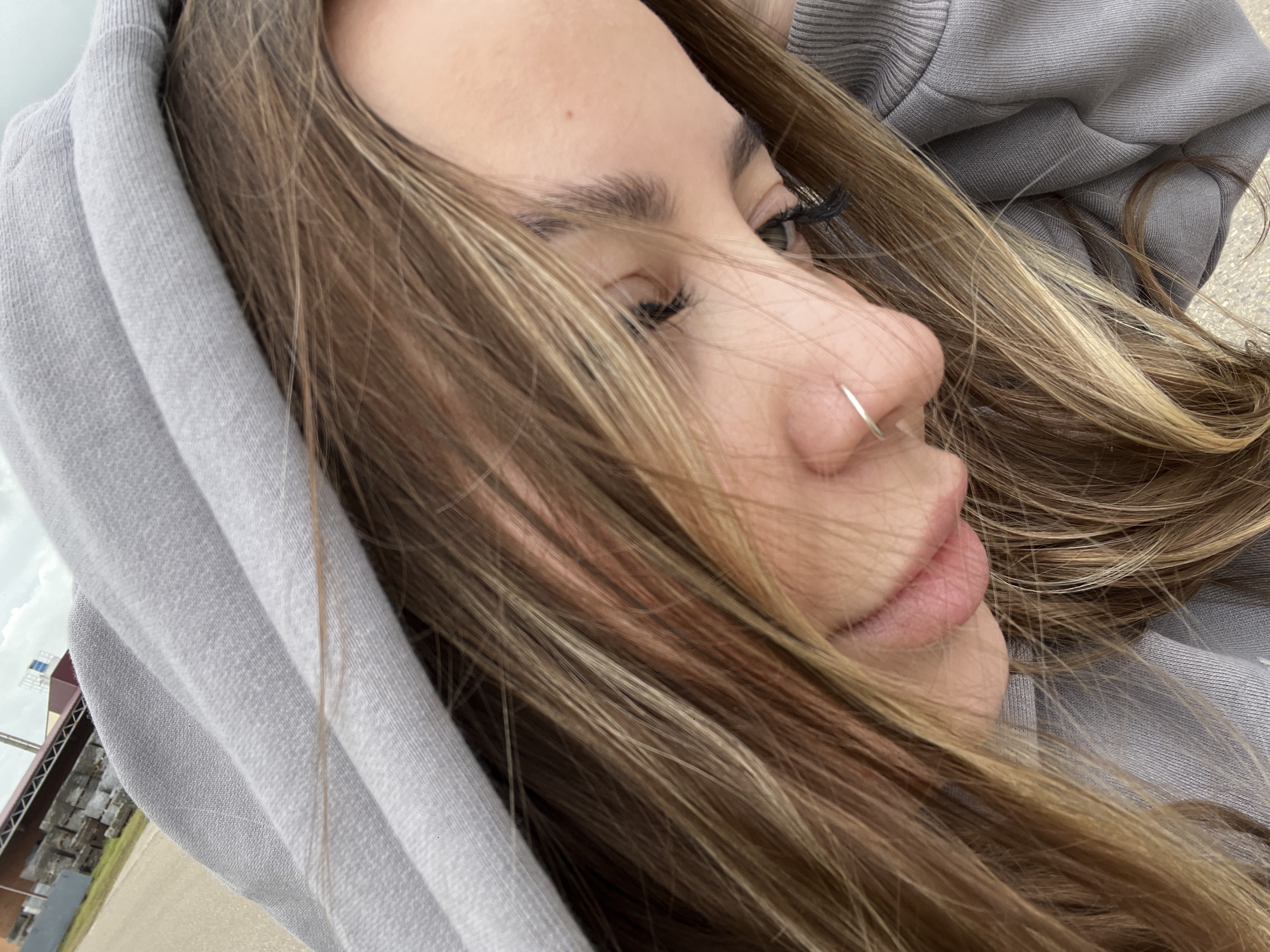 A close-up, angled shot shows a young woman with light brown hair highlighted with blonde streaks. She is wearing a grey hooded sweatshirt, with the hood partially covering her head. Her face is tilted slightly downward, and she is looking to the side. A small silver hoop is visible in her left nostril. She is wearing false eyelashes. The background shows a building and some vegetation, and is potentially in Novopokrovskaya, Russia.