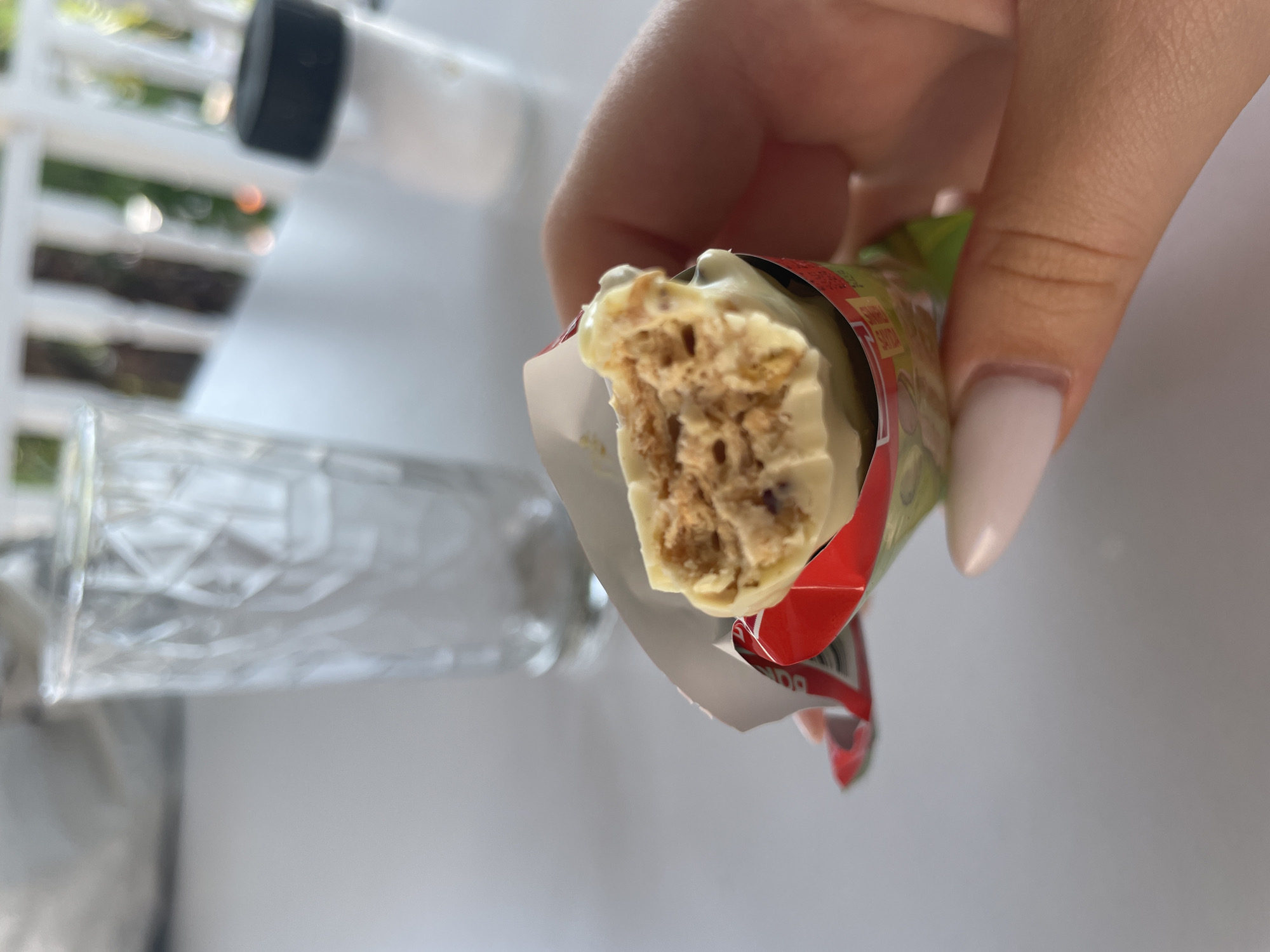 A person's hand, with neatly manicured light pink nails, holds a partially unwrapped snack bar. The snack bar appears to have a white chocolate coating and a crunchy, multi-textured interior with visible brown pieces. The wrapper is red and green. In the background, a clear glass with a textured pattern is visible, along with a salt shaker with a black lid. The setting appears to be outdoors, possibly on a table on a balcony or patio, with a blurred background of white railings and greenery, suggesting a location in Skala Avdiron, Greece.