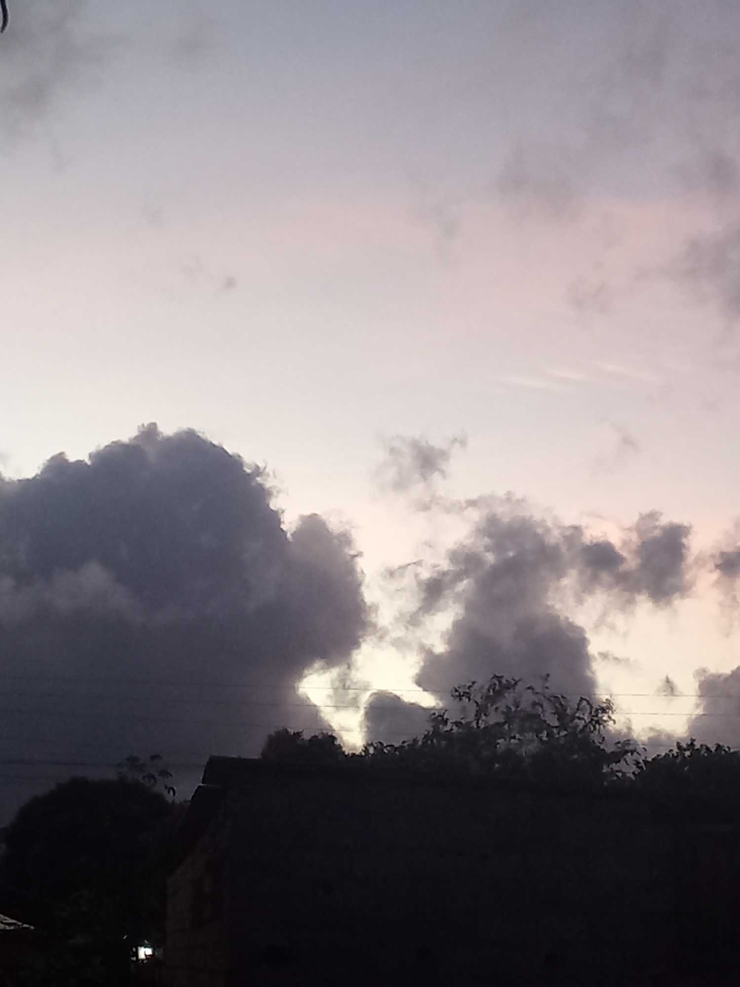 The image depicts an outdoor scene at twilight or dawn in Juan Griego, Venezuela. The sky occupies the majority of the frame, displaying a gradient from a pale purplish-pink and light grey in the upper sections to darker, volumetric grey and black cumulus clouds in the lower two-thirds. Multiple horizontal utility lines are visible across the mid-sky, intersecting with the cloud formations. Below the horizon line, the landscape is rendered in dark silhouette, consisting primarily of dense tree foliage and several indistinct building structures. One prominent structure, located to the right of the lower frame, features a distinct, possibly angled or tiered, roofline. A small, bright, rectangular artificial light source is discernible at ground level in the lower-left quadrant, partially obscured by foreground elements. No individuals, animals, or discernible activities are present.