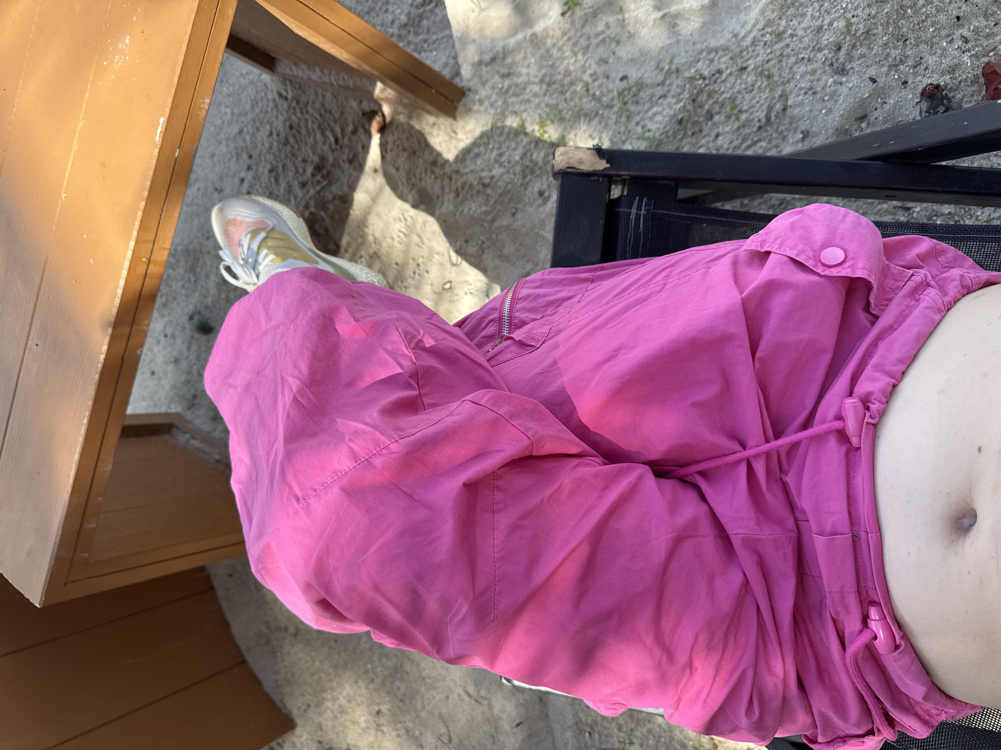 The media file captures a close-up, overhead view of a person's midsection and legs, seated outdoors on a textured surface. The person is wearing bright pink, loose-fitting pants with a drawstring waist, a zipper closure, and a small pink button on a flap. A white and pink sneaker is visible on the left foot. The background consists of sand, gravel, and some sparse greenery, suggesting an outdoor setting. To the left, a portion of a wooden table with a light brown finish is visible, with sunlight casting shadows. To the right, the edge of a black chair or lounge is present, with some visible wear and tear.
The lighting suggests it is daytime, likely outdoors. The shadows indicate the sun is at an angle, possibly late morning or afternoon. There are no discernible emotions conveyed due to the focus on clothing and setting. No text is visible in the image. The overall impression is one of casual relaxation in a natural or semi-natural outdoor environment. The setting appears to be Baan Plai Laem, Thailand, as per the provided location context.