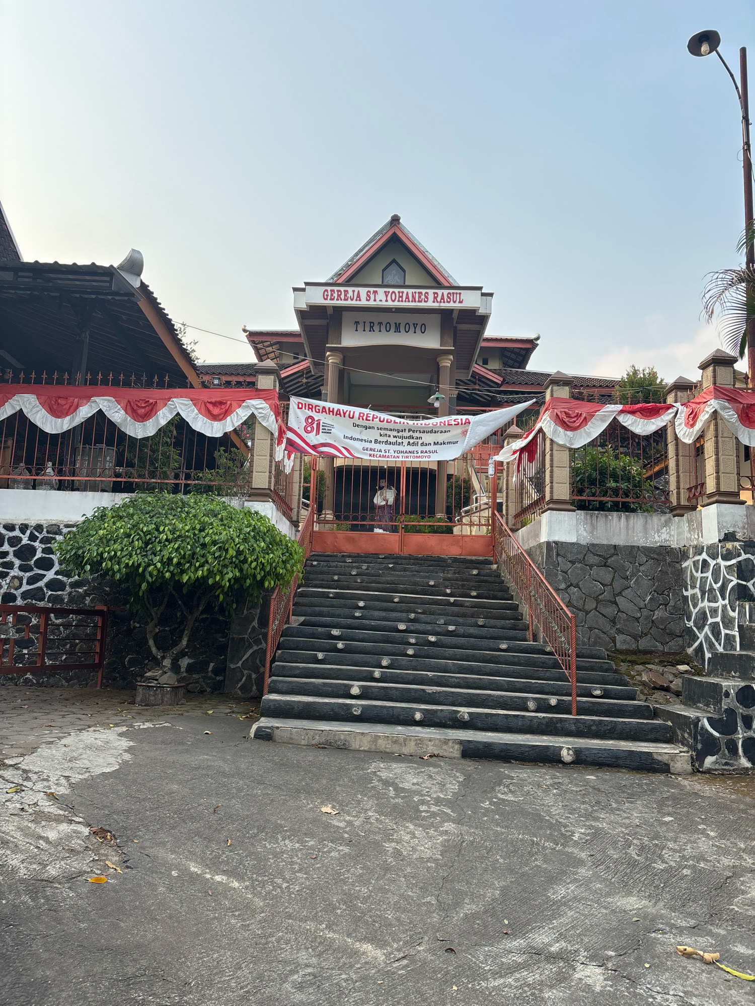 This is a photograph of the Gereja St. Yohanes Rasul church in Tirtomoyo, Indonesia. The church building has a prominent facade with the name "GEREJA ST. YOHANES RASUL" and "TIRTOMOYO" displayed above the entrance. A banner celebrating "DIRGAHAYU REPUBLIK INDONESIA" with the number "81" is hung across the front of the building, partially obscured by decorative red and white fabric draped along a fence.
In front of the church is a wide set of stairs, each step illuminated by a small light. A red metal railing runs alongside the stairs. The stairs lead up to a gated entrance where a person, possibly a visitor or attendee, is visible standing just inside.
The church is situated on a raised stone-and-concrete foundation, with a manicured green tree on the left side of the stairs. The ground in front of the stairs is a paved asphalt area with some scattered leaves. A tall lamppost stands to the right of the building. The sky is clear and bright, suggesting it is daytime. The overall scene depicts a place of worship decorated for a national celebration.