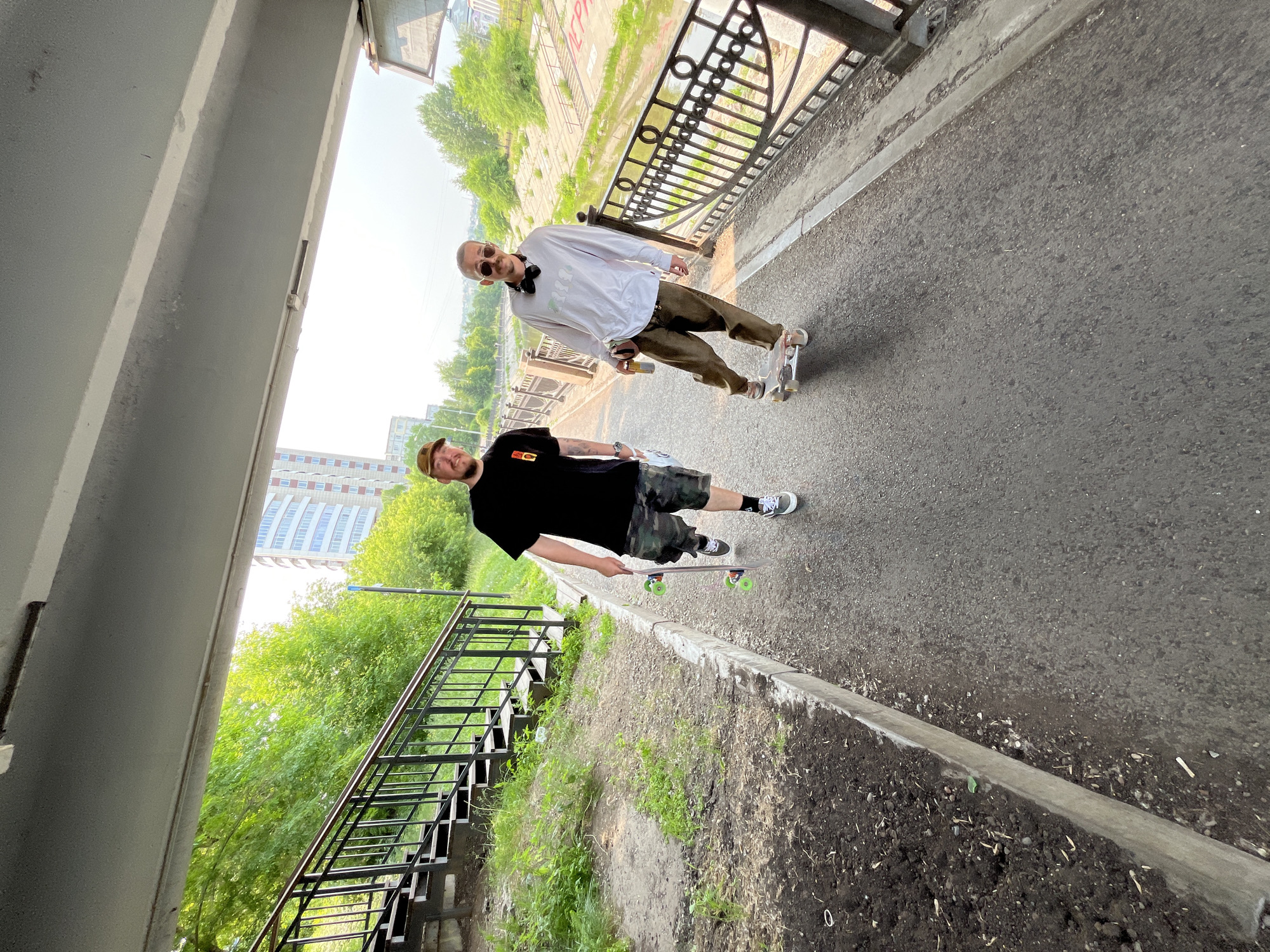 Two men are walking under a pedestrian bridge in the city of Krasnoyarsk, Russia. The man on the left is wearing a black t-shirt, camouflage shorts, and a baseball cap. He is holding a skateboard. The man on the right is wearing a white long-sleeved shirt, brown pants, and sunglasses. He is standing on a skateboard and holding a can. The bridge is made of concrete. To the left of the path are stairs and vegetation. To the right of the path is a railing and a river. The sky is overcast.