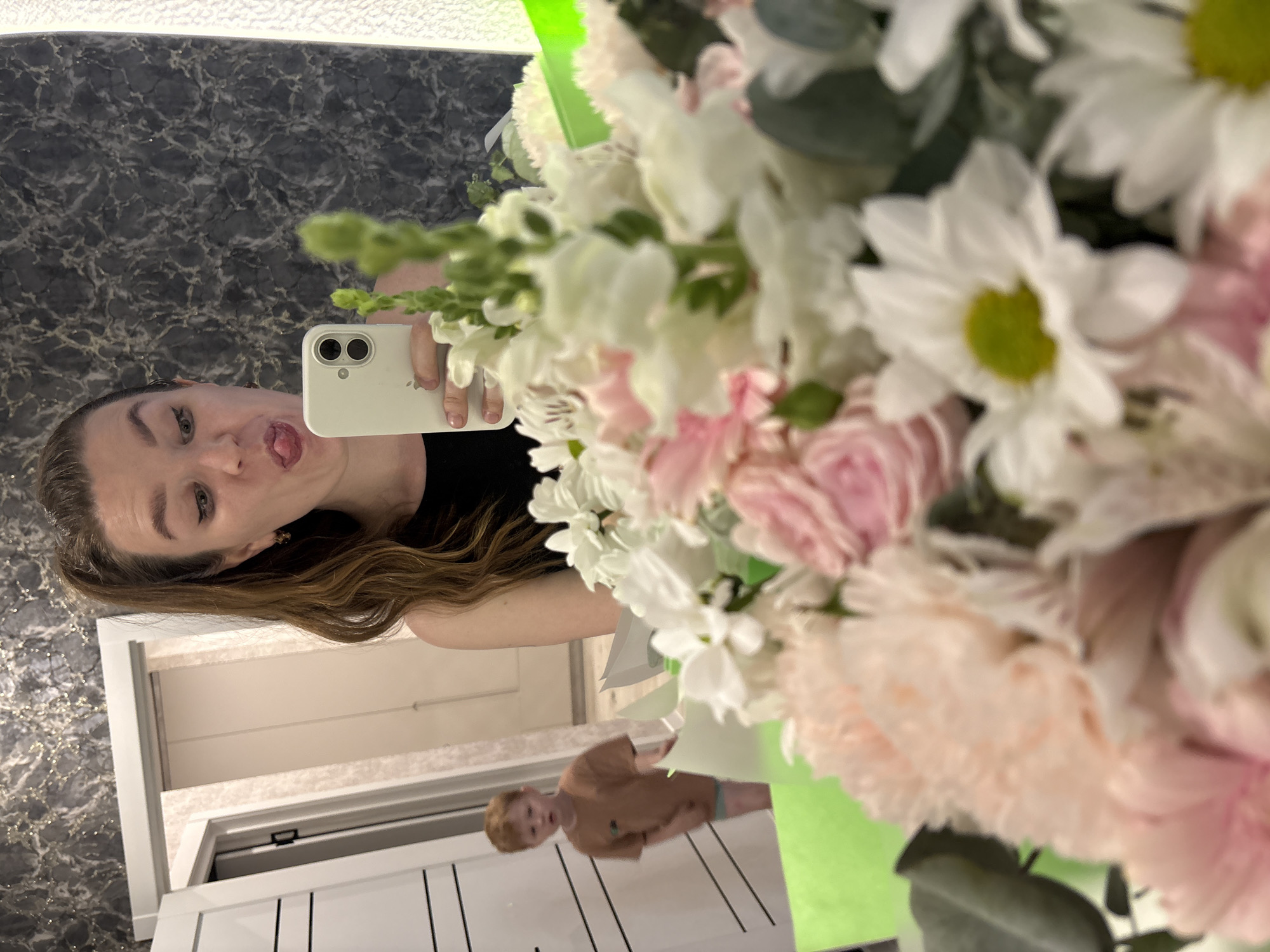 A woman is taking a selfie in a mirror. She is holding a white smartphone in her right hand and sticking her tongue out at the camera. She has long, wavy brown hair and is wearing a black top. In front of her is a large bouquet of white and pink flowers. The flowers appear to be daisies, roses, carnations, and snapdragons. In the mirror reflection, a child is visible, wearing a brown shirt. The background shows a dark marble patterned wall, a white door, and a glimpse of the interior of a room. Based on the information available, I cannot determine if the location is in Izhevsk, Russia, as there are no visible indicators to confirm it.