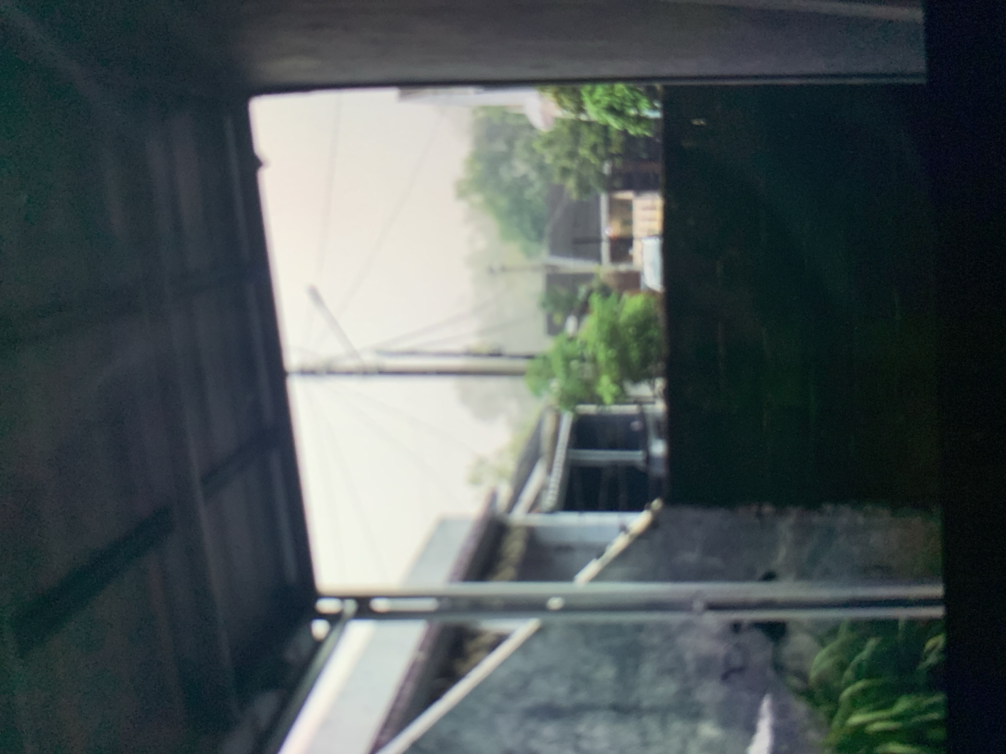 The image is a slightly out-of-focus, vertical view from a covered outdoor space, possibly a porch or carport, looking out onto a residential street in Ciamis, Indonesia. The foreground is dominated by the dark interior of the covered area, with a dark ceiling and the edge of a concrete wall on the right. Through the opening, a residential scene is visible. On the left, a light-colored wall of a building is prominent, with a dark, angled roofline above it. Further into the scene, more buildings with sloped roofs are visible, some appearing to be single-story houses with open verandas supported by columns. Trees with lush green foliage are scattered throughout the background, suggesting a tropical or subtropical environment. A utility pole with wires is also visible in the distance. The lighting suggests it is daytime, possibly overcast or hazy, as the light is diffused and the overall atmosphere is muted. There are no people or animals clearly visible in the frame. The scene depicts a typical residential neighborhood in a city like Ciamis, characterized by modest homes and abundant greenery.