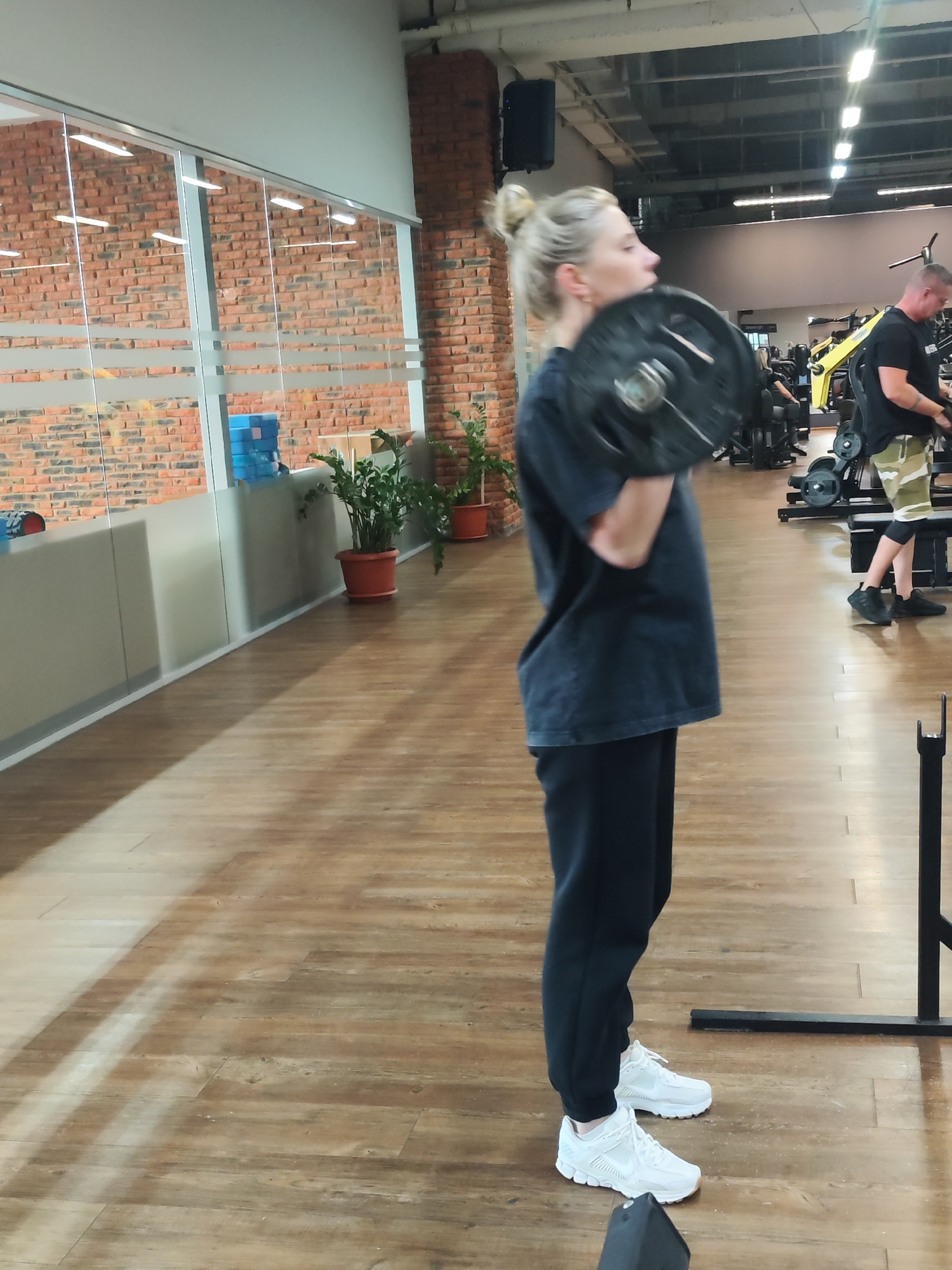 A woman with blonde hair pulled up in a bun is at a gym in Chaikovsky, Russia. She is wearing a dark blue t-shirt, black sweatpants, and white sneakers. She is holding a weight plate close to her chest. In the background, a man is standing near workout equipment. The gym has a brick wall with windows, plants in pots, and wooden floors.