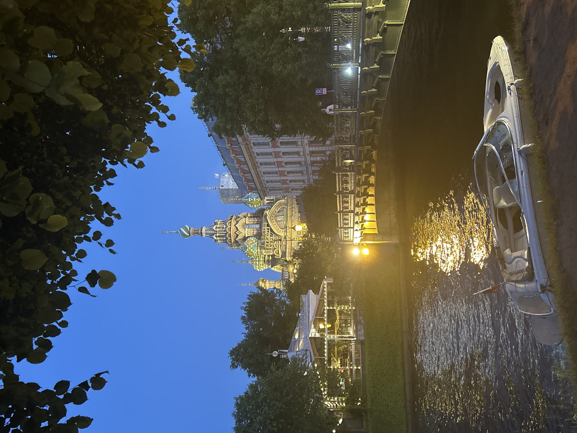A nighttime scene unfolds under a deep blue sky, framed by the dark silhouettes of tree branches laden with leaves. In the distance, a majestic, elaborately decorated church with a vibrant green and blue onion dome dominates the skyline. Adjacent to it stands a multi-story building with a red-tiled roof and pale walls, punctuated by numerous windows.
In the foreground, a calm body of water reflects the warm glow of lights. A sleek, white motorboat, bearing the number "513" on its side and a small flag with red, white, and blue stripes, rests partially on the grassy bank. A decorative bridge with intricate metalwork spans the water, illuminated by warm lights, and a building with a glass roof and twinkling string lights sits on the embankment. A single street lamp with a classical design stands near the bridge. Two individuals are visible on the bridge, one standing and looking towards the scene, and another further down. The overall atmosphere is serene and picturesque, capturing the charm of a city at twilight.