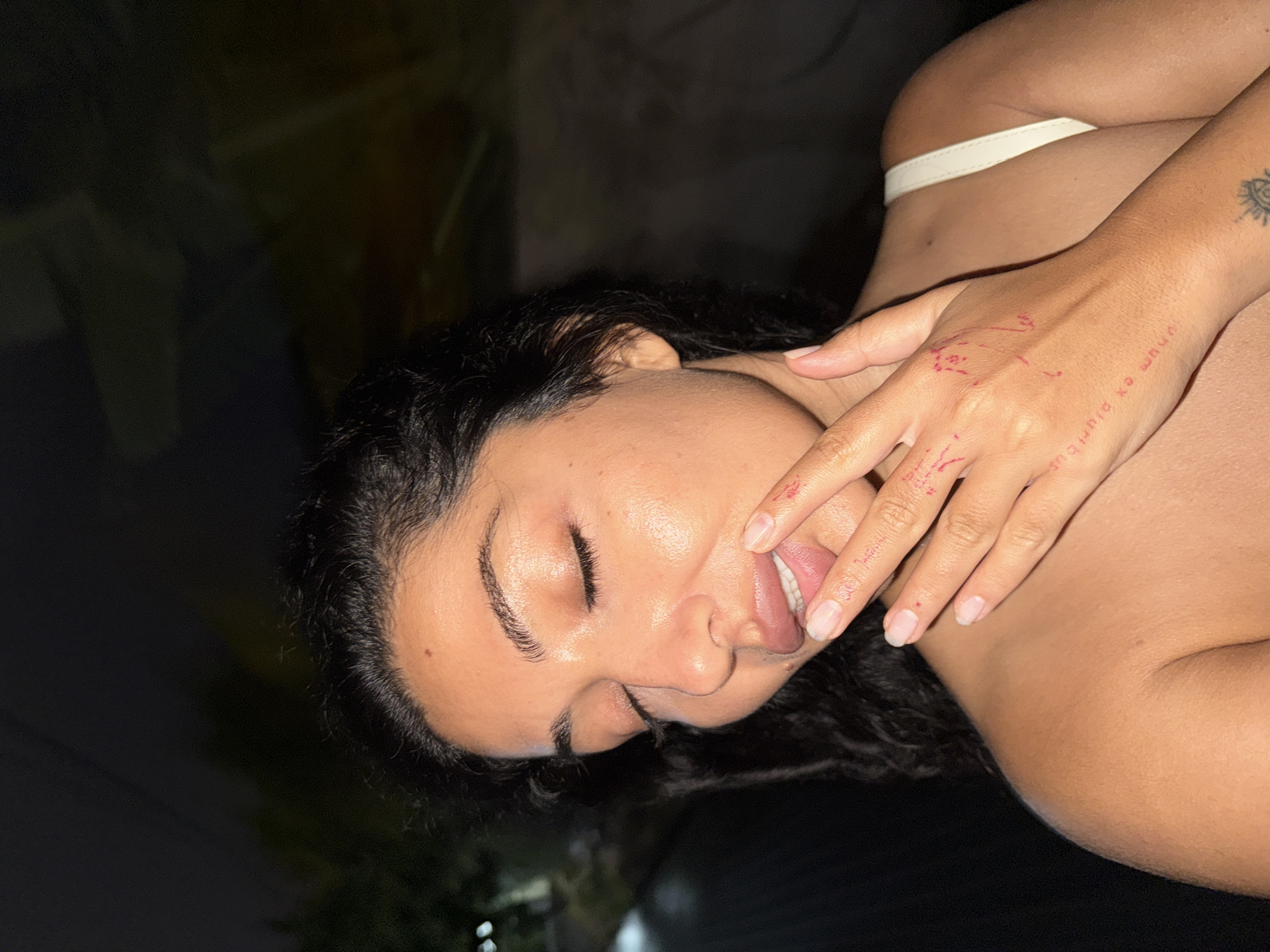 A close-up, rotated shot captures the upper body and head of a person, likely a woman, with dark, wavy hair and tan skin. The individual is lying on their side against a dark background, suggesting a nighttime setting or a dimly lit interior. The context indicates the location is North Kuta, Indonesia.
The person's eyes are mostly closed, and their right hand is positioned over their mouth, with fingers slightly entering the mouth and the tongue partially visible. This creates an intimate or candid expression. Red markings, resembling temporary tattoos or writing, are visible on the back of the right hand. The Latin phrase "unum ex pluribus" ("one out of many") is clearly readable on the hand, alongside other red patterns and dots on the fingers and palm area. A small, stylized sun or eye tattoo is visible on the person's right forearm.
A white strap, possibly from underwear or a bra, is visible around the person's midsection. The lighting is low, emphasizing the textures of the skin and hair. The person's nails appear natural and unpolished. The overall mood of the image is intimate and relaxed.