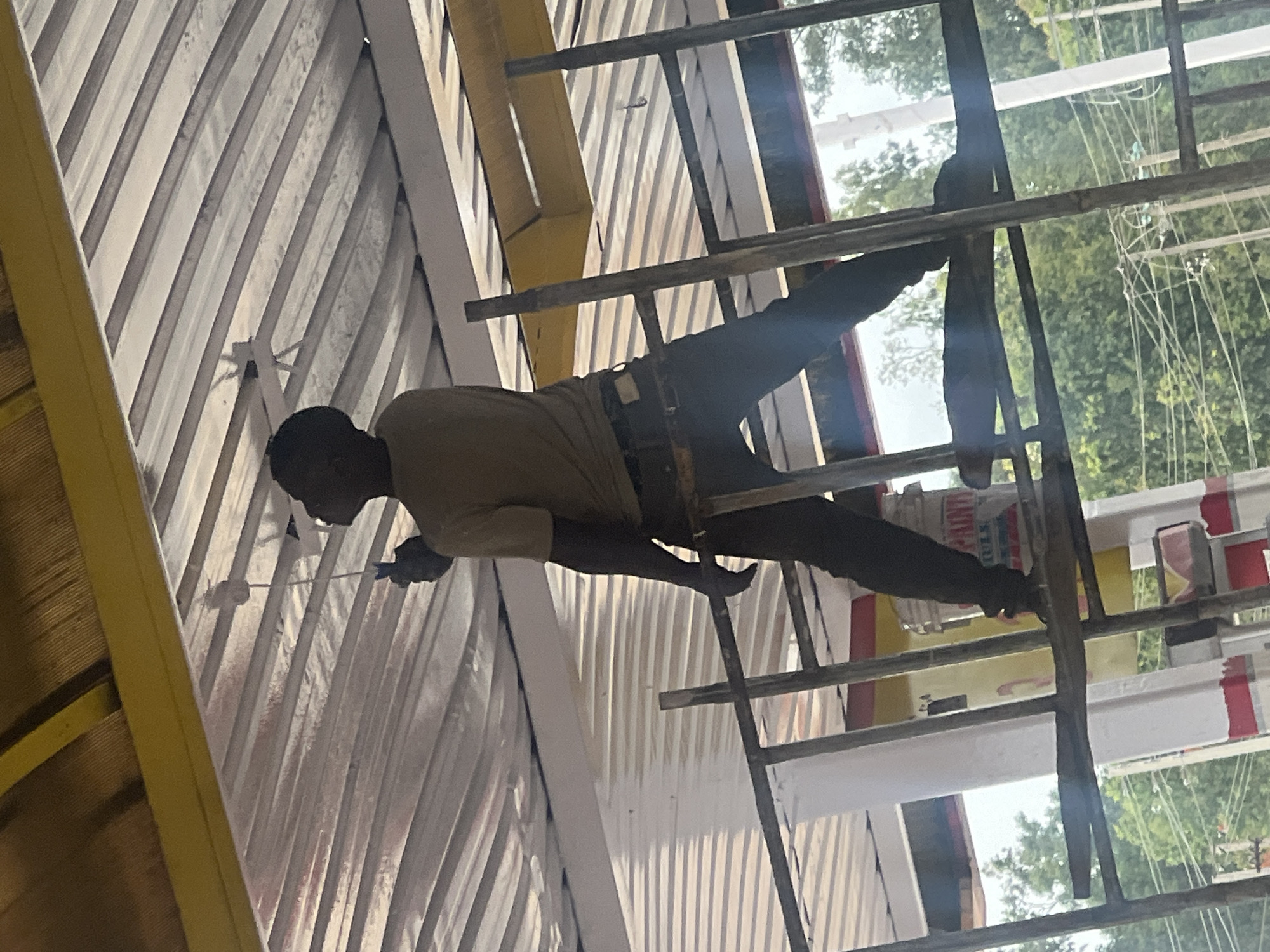 A construction worker paints a tall structure in Gura Top, Nigeria. He stands on scaffolding, clad in dark pants and a tan shirt with a belt. The structure consists of many angled, parallel white slats. The worker holds a paint brush in his left hand. The scaffolding is made of metal. Trees are visible in the background.
