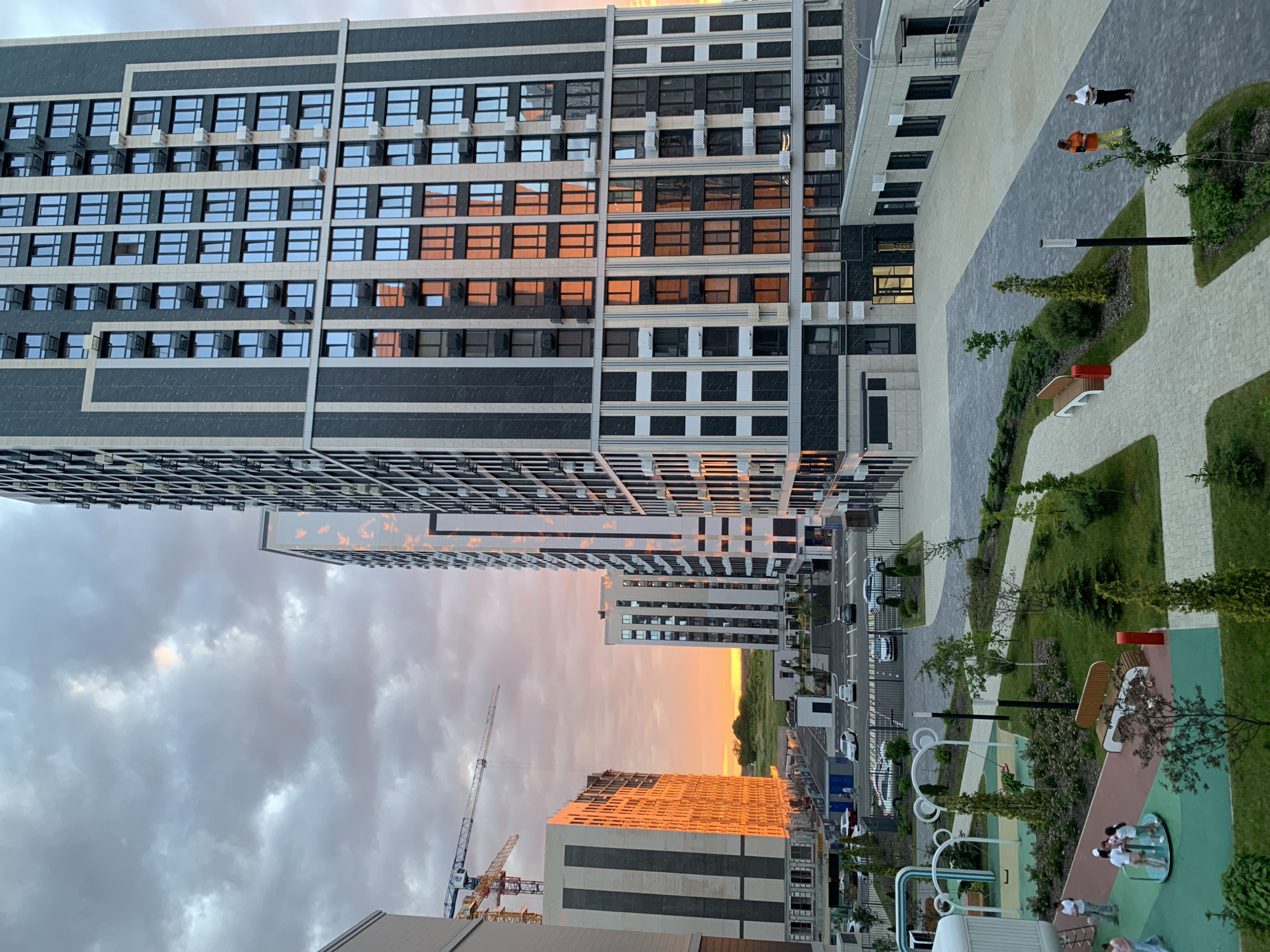 The image depicts an outdoor scene in a modern urban development, likely a residential complex. The time of day is sunset, as evidenced by the warm, orange glow illuminating the sky and reflecting on the facades of buildings. Dominating the foreground and right side of the image are tall, contemporary apartment buildings with a dark gray and beige facade, featuring numerous rectangular windows.
On the left side of the image, another large building is under construction, with its windows reflecting the sunset intensely. In the background, a bright orange sunset stretches across the horizon, with some trees and greenery visible. Construction cranes are also present in the distance, indicating ongoing development.
In the lower part of the image, there is a playground area with colorful equipment, including swings and a merry-go-round. Several children are playing on the merry-go-round. A pathway made of interlocking pavers winds through a landscaped area with grass, bushes, and young trees. Benches are placed along the path.
Further back, a parking lot is visible with several cars parked. Two women are standing and conversing near a paved courtyard area, in front of another low-rise building. The overall impression is one of a new, developing residential area at dusk.