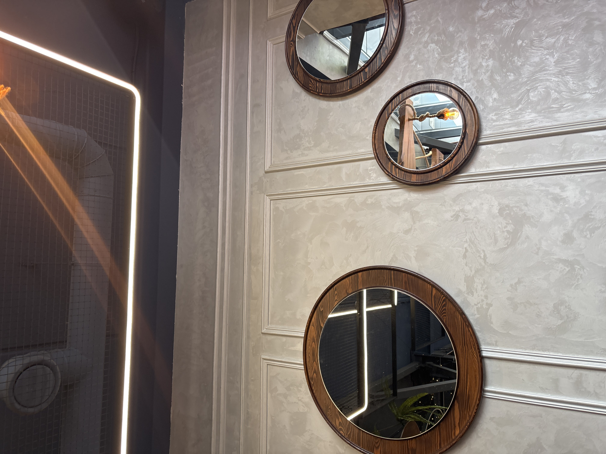 The image depicts an interior scene featuring three oval mirrors mounted on a decorative textured wall. The mirrors have wooden frames and reflect various elements of the room, including lighting fixtures and structural components of the ceiling. The wall itself has a light, mottled texture, with vertical paneling details. Above the wall, a dark ceiling with a grid pattern is visible, along with recessed lighting and a ventilation duct. The overall impression is one of a modern, possibly commercial, interior space. This image was taken in Naberezhnye Chelny, Russia.