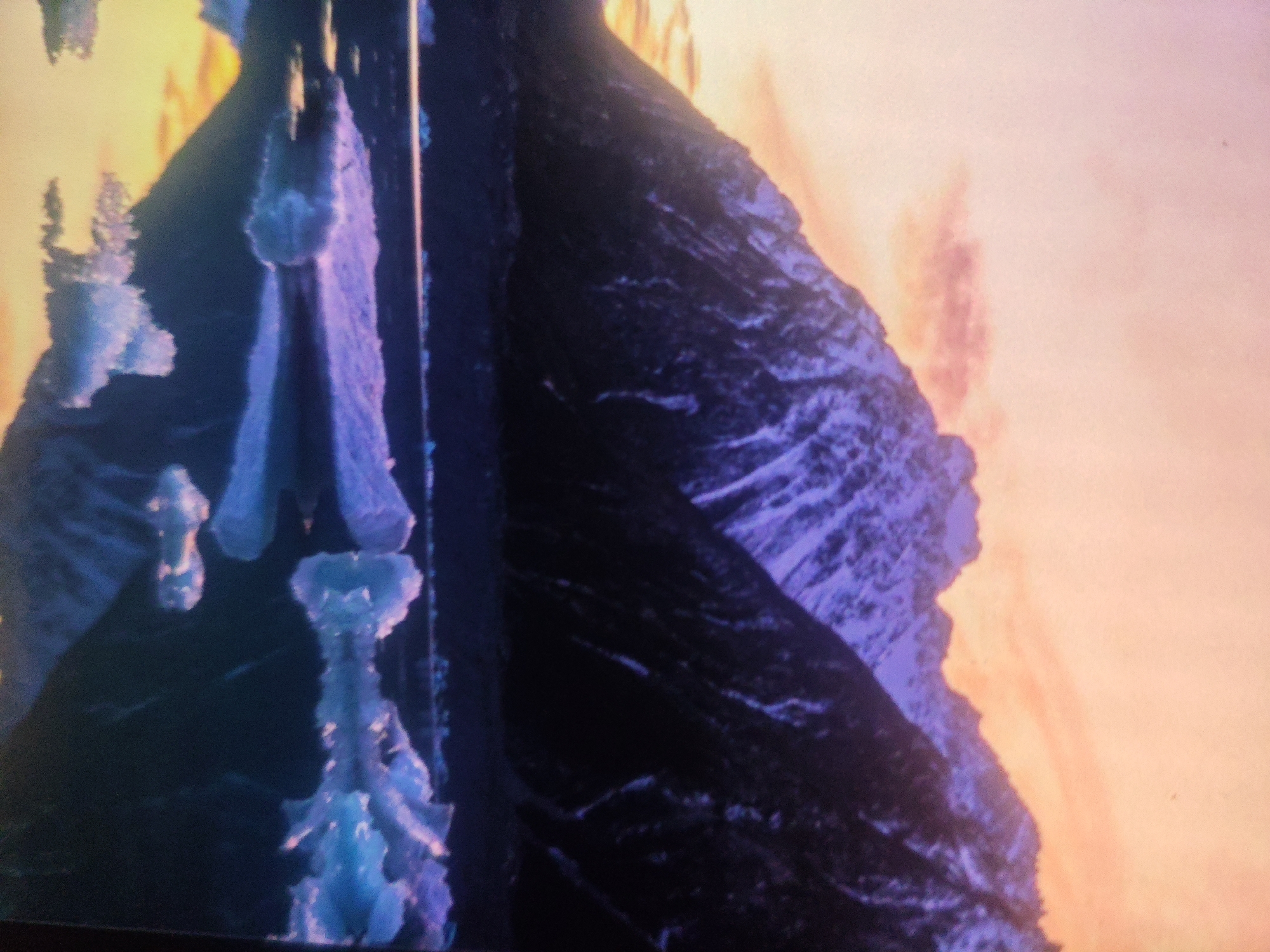 The image shows a close-up view of an abstract, potentially digital or artistic rendering with a focus on texture and color. On the left side, there are vertical structures with a crystalline or icy appearance, rendered in shades of bright blue and purple, with some lighter, almost white highlights. These structures have a somewhat organic, layered, or faceted texture.
To the right of these blue/purple structures, there is a dark, textured surface that appears mountainous or rocky, predominantly in deep blues and purples, with lighter streaks suggesting snow or texture. This dark area dominates the right half of the image and extends upwards.
The background on the far right transitions into a soft, luminous gradient of colors, ranging from pale orange to pinkish-yellow, creating a hazy or atmospheric effect. This background has subtle vertical streaks of a slightly darker pink or orange.
There are no discernible people, animals, or recognizable objects from a typical urban environment like Armavir, Russia. The scene is abstract and does not depict any specific location in the city. The overall impression is one of an otherworldly or digitally created landscape with a strong emphasis on textural detail and vibrant, contrasting colors.