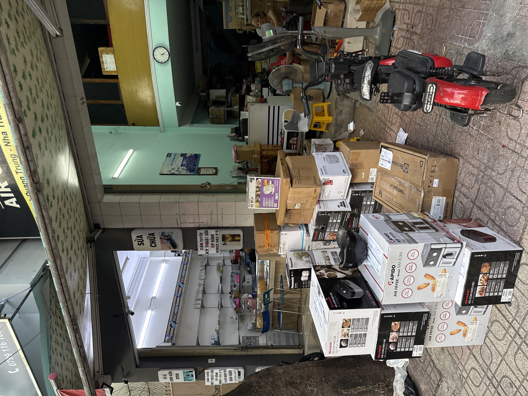 The image depicts an outdoor storefront in Nha Trang, Vietnam, likely a small electronics or appliance shop. A variety of boxed goods, primarily rice cookers and air fryers from brands like Sharp and Rapido, are stacked haphazardly on the ground in front of the entrance. A red electric scooter is parked to the right of the merchandise. In the background, the shop interior is visible, featuring shelves stocked with more products and a counter. A woman is seated inside the shop on the right. A clock on the wall indicates the time is approximately 11:05. The storefront has awnings and signage, including Vietnamese text indicating "Sửa Chữa" (Repair) and "Cần Tuyển Kỹ Thuật Sửa" (Hiring Repair Technicians). The overall scene suggests a busy retail environment with goods displayed both inside and outside the shop.