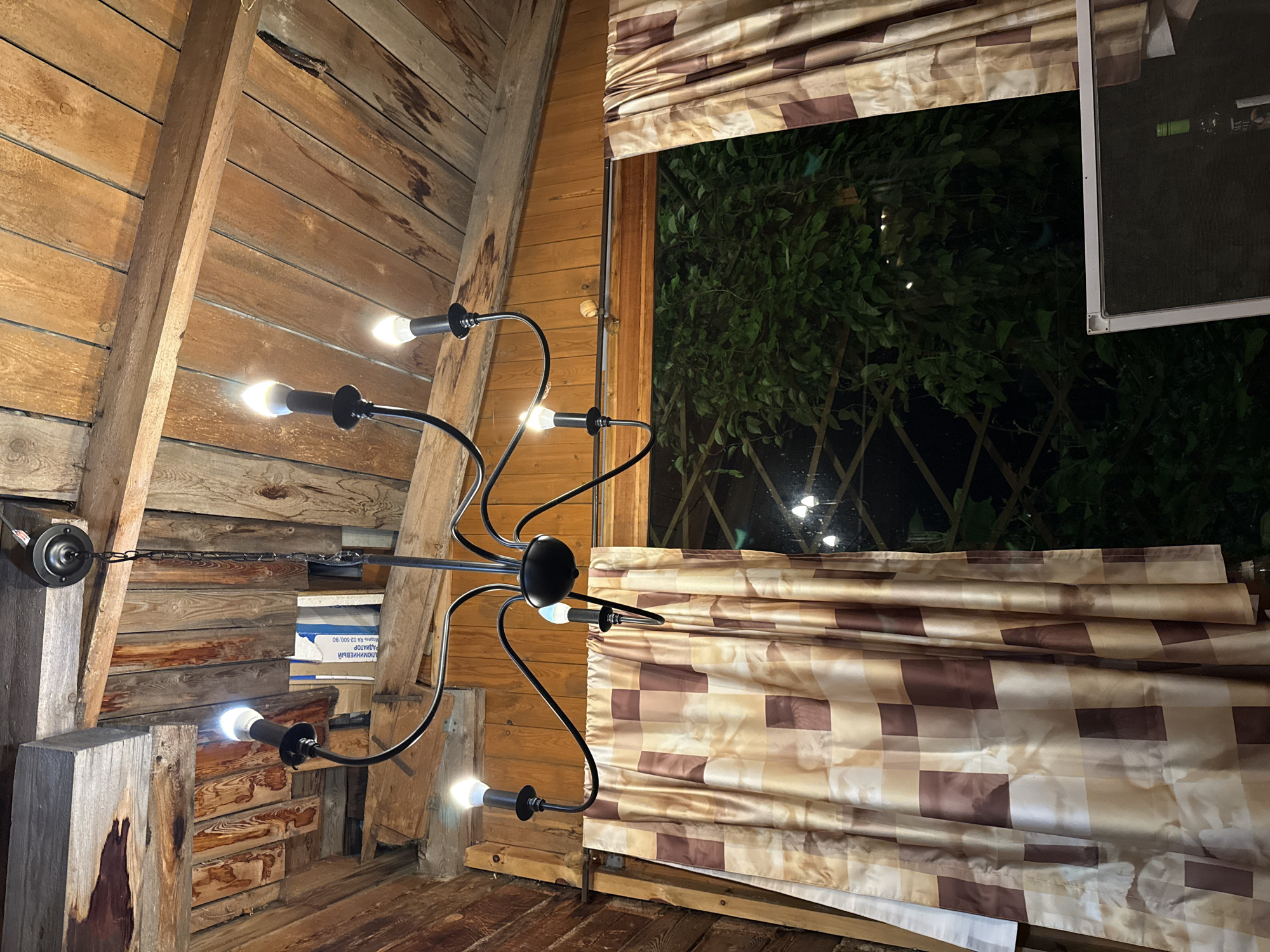 A black chandelier with six bulbs hangs from a wooden ceiling. The chandelier is centered in the frame, with its arms extending outwards. The bulbs are lit, casting a warm glow. To the left of the chandelier, a brown and beige checkered curtain is drawn across a window. The window looks out onto a dark, leafy exterior with some faint lights visible in the distance. The walls and ceiling are made of dark wood planks. The scene appears to be indoors, possibly in a rustic or cabin-like setting. There are no people visible in the image. The location is not specified in the image itself but is given as Sovetskiy, Russia.