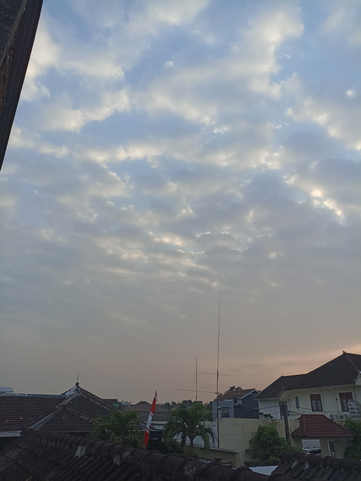 This is a photograph taken from a rooftop perspective, looking upwards towards a cloudy sky. The sky is filled with a pattern of cumulus clouds, with patches of sunlight breaking through, suggesting it might be early morning or late afternoon. In the foreground, several tiled rooftops are visible, with a darker, more textured rooftop in the very bottom of the frame. Among the rooftops, there are glimpses of greenery from palm trees. To the right, a white building with a dark-tiled roof is prominent, featuring windows and what appears to be an air conditioning unit. A small red and white flag, likely the Indonesian flag, is attached to a pole on one of the rooftops. In the middle ground, several communication towers or antennas are visible, extending above the buildings. The background shows more houses and trees, with the sky transitioning to a lighter hue near the horizon. The overall impression is a residential area in a tropical climate, possibly the city of Sumberejo, Indonesia, during a time of diffused daylight.