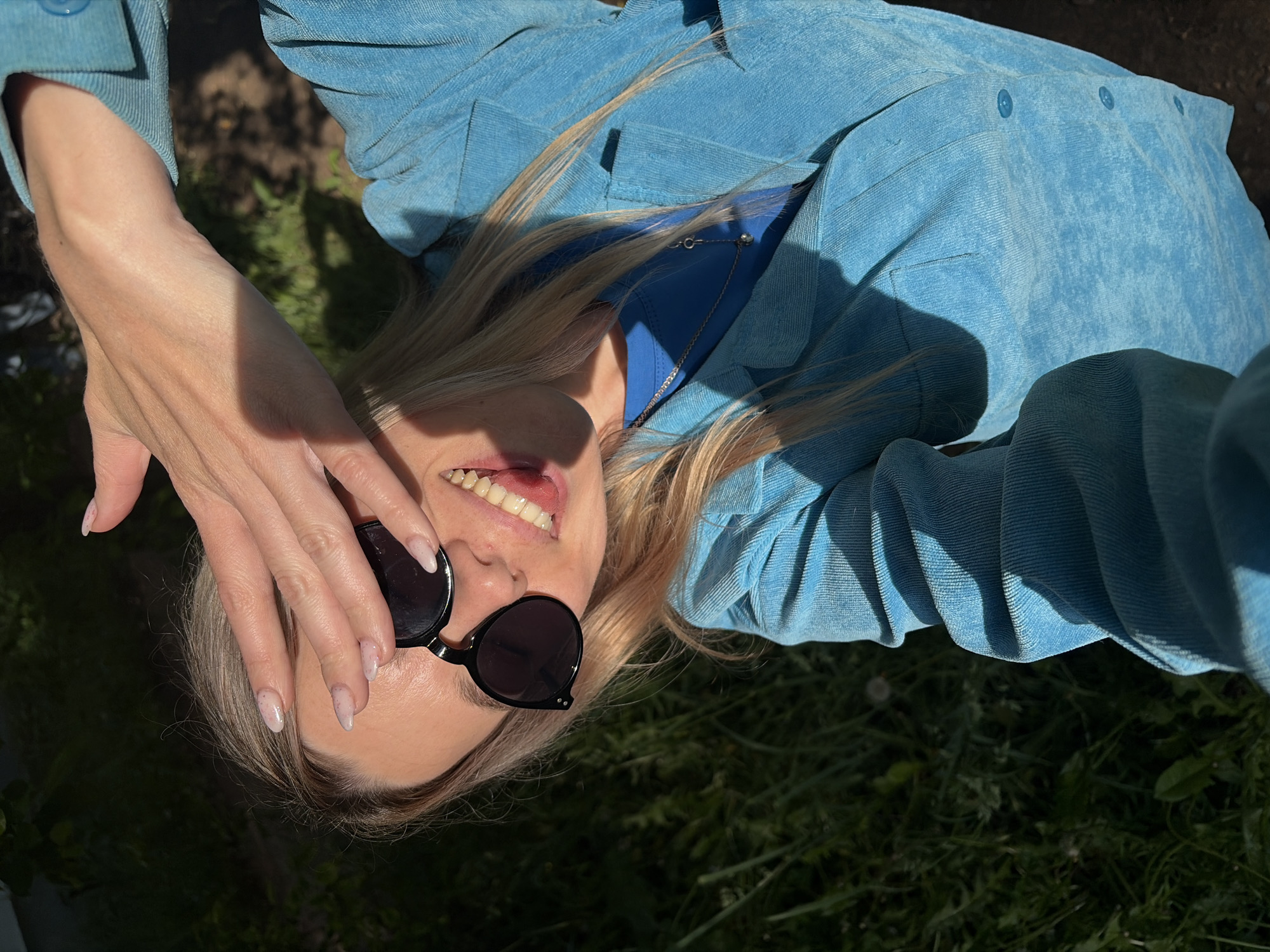 The image shows a close-up, high-angle shot of a person, likely a woman, with blonde hair, lying on a patch of grass in Chaikovsky, Russia. She is wearing a light blue, textured jacket, possibly corduroy, over a plain blue top, and a silver chain necklace is visible around her neck. Her left hand, with light-colored manicured nails, is raised to cover part of her forehead, casting a shadow, and also rests near her black, round sunglasses. The person is smiling broadly, revealing white teeth and a partially visible tongue, conveying a sense of joy, playfulness, or amusement. Strong, direct sunlight creates sharp shadows on her face, arm, and clothing, emphasizing the textures of her attire and the surrounding environment. The background consists of vibrant green grass mixed with some darker earth patches, indicating an outdoor setting during a sunny day. No text is visible in the image.