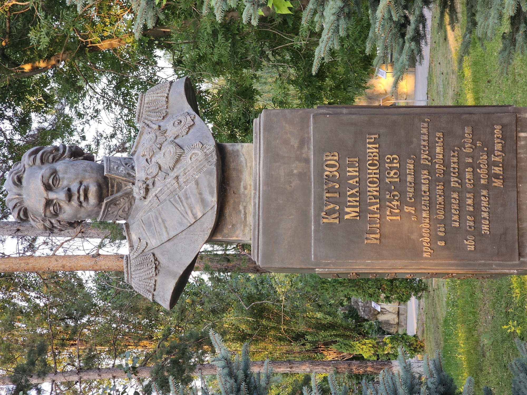 A bronze bust of Mikhail Kutuzov is mounted on a stone pedestal in a park in Cherkessk, Russia. The bust depicts Kutuzov from the chest up, with a stern expression and a military uniform adorned with medals. The pedestal has an inscription in Russian, detailing his name, dates of life (1745-1813), and a quote attributed to him. The surrounding environment consists of lush green trees, grass, and some sparse vegetation. The sun is casting warm light on the scene, creating a peaceful atmosphere.