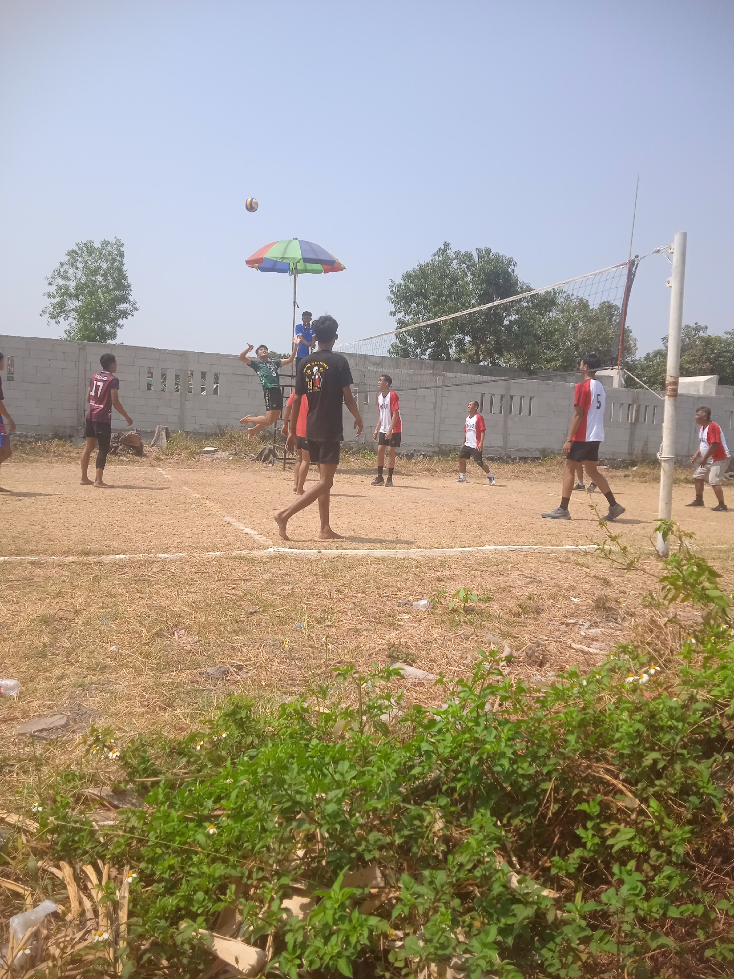 In Klodran, Indonesia, a volleyball game is underway on a dirt court under a clear sky. The court is marked with white lines and surrounded by dry grass and sparse vegetation. A volleyball net stretches across the middle of the court, supported by white poles. Several individuals are actively participating in the game. One player in a green jersey is mid-air, about to hit the ball, while another player in a black t-shirt stands nearby. Other players in red and white jerseys are positioned on the court, some facing the net and others observing the play. A multi-colored umbrella is set up behind the net, possibly for shade. A concrete wall with openings runs along the background, and trees are visible in the distance.