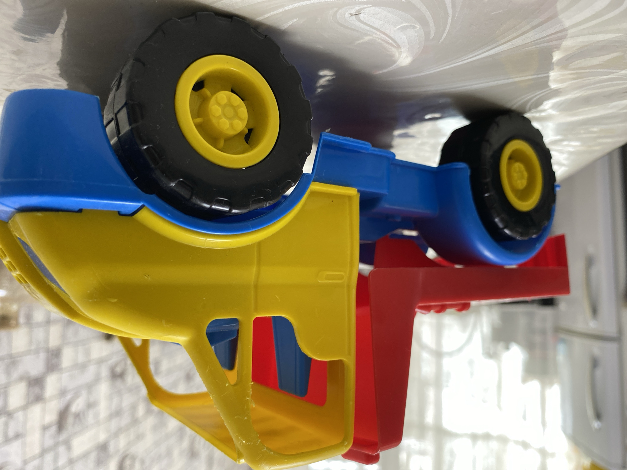 A brightly colored plastic toy truck, predominantly yellow, blue, and red, sits on a table with a patterned surface. The truck is a model of a towing vehicle, featuring a yellow cab with blue interior seats and a red rear section. It has large black tires with yellow rims. In the background, a white refrigerator is visible, along with a window with white curtains, suggesting an indoor setting. The overall scene is likely in a residential area within Бескөл ауылдық округі, Kazakhstan, given the context. There are no people or animals present in the image.