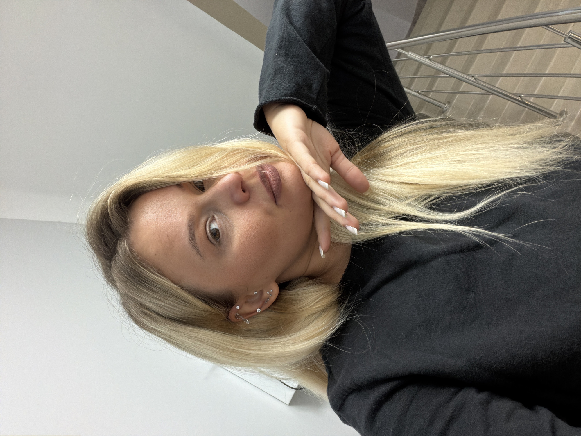 A fair-skinned person with blonde hair and multiple ear piercings is shown in a selfie-style portrait. The person is wearing a black long-sleeved garment. The background features a white wall and a metal railing, possibly indicating a staircase or balcony. There is no information to verify this image was taken in Sarapul, Russia.