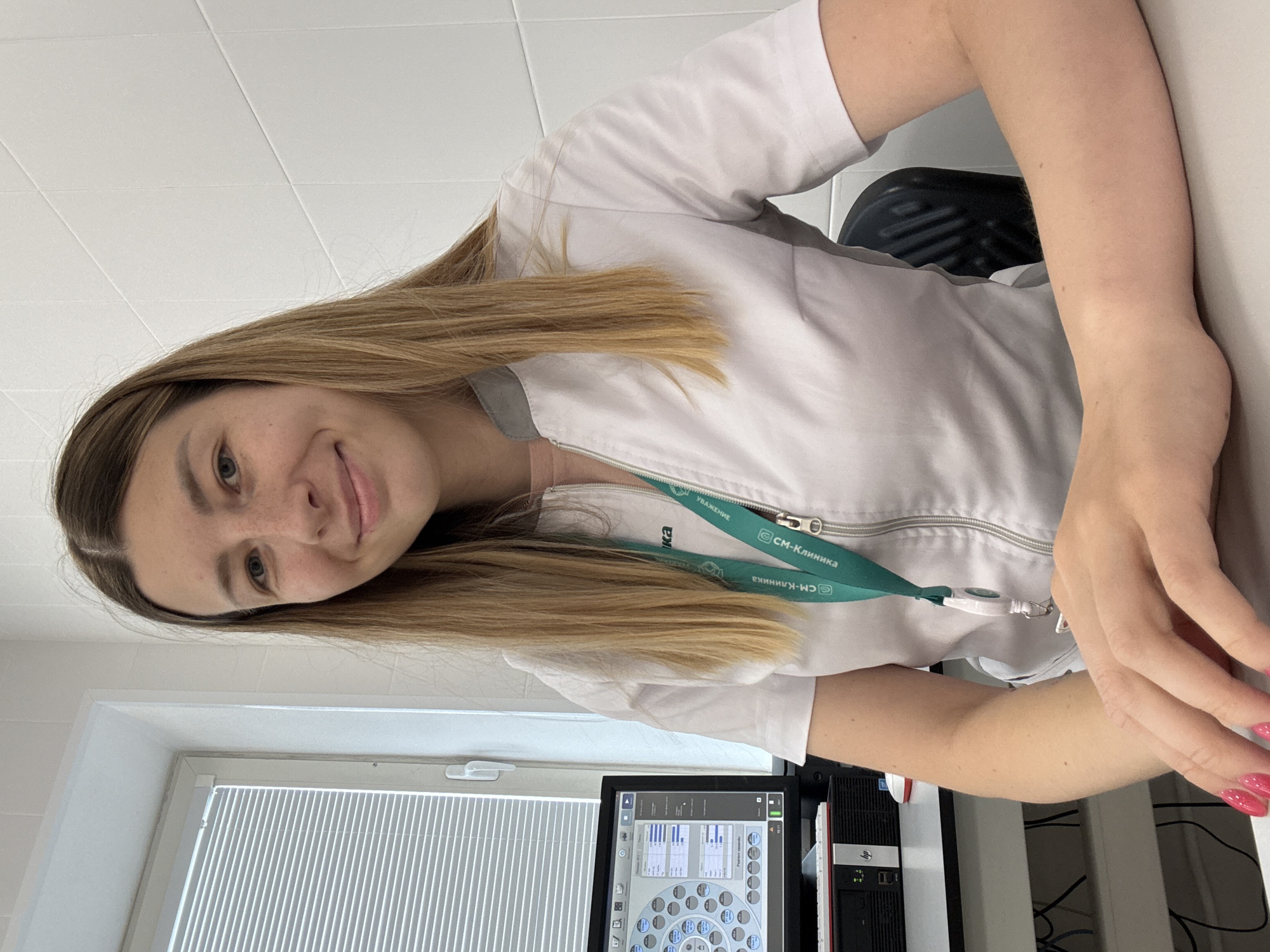 A young adult female with blonde hair and freckles is sitting at a desk in a brightly lit room. She is wearing a white lab coat over a pink shirt and a green lanyard with a badge. She is looking at the camera and smiling. In the background, there is a computer monitor displaying medical data, a window with blinds, and a tiled wall. The scene appears to be in a clinic or laboratory setting.