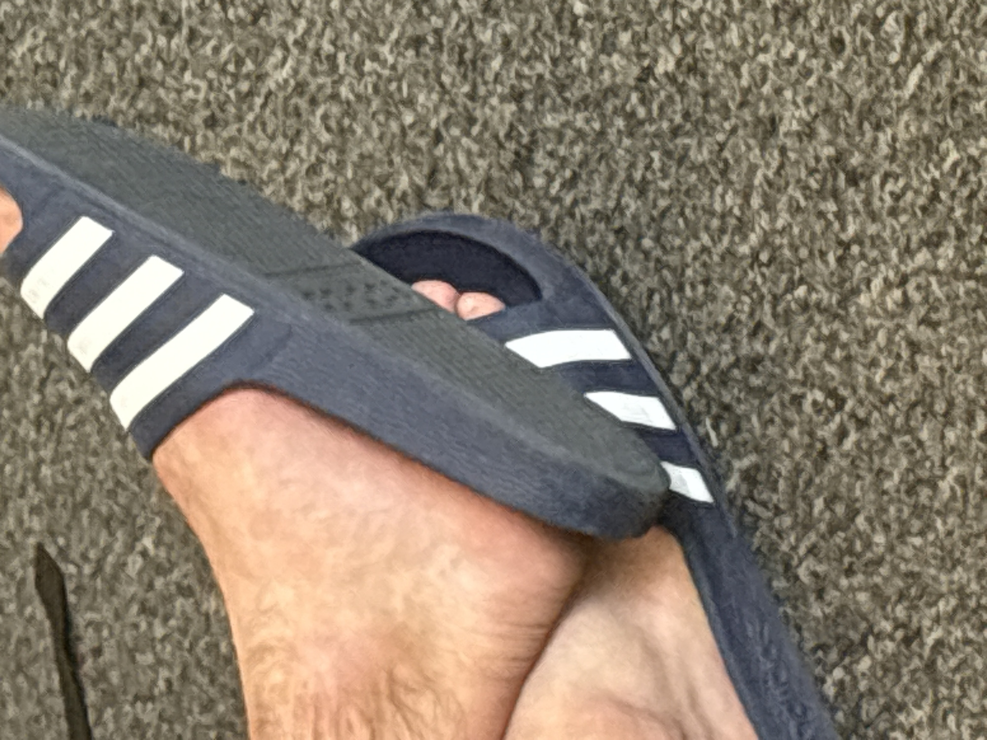 A close-up shot shows a person's feet wearing navy blue Adidas slides. The slides have three white stripes on the strap. The person's feet are resting on a textured, grey carpet. The scene is lit by natural light, casting soft shadows. The image is a still shot, with no activity or event depicted. The focus is on the footwear and the texture of the carpet. There is no visible text.