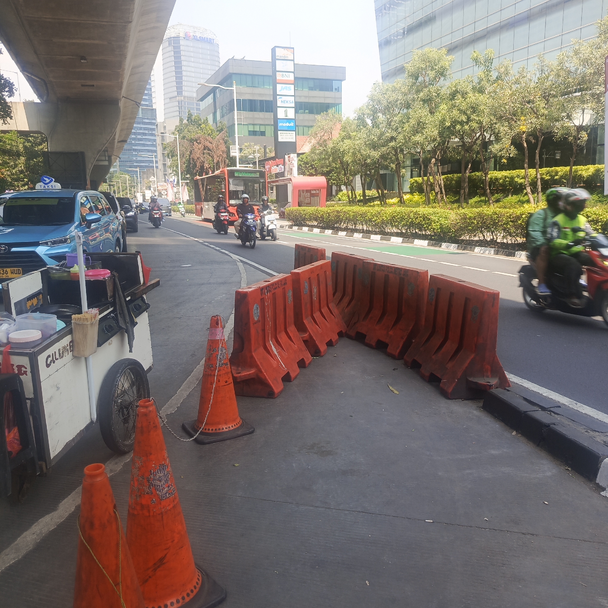 The image shows a street scene in South Jakarta, Indonesia, likely during the daytime with bright sunlight. In the foreground, an orange street food cart is parked on the left side of the road, displaying various containers and utensils. Two orange traffic cones are placed on the road near the cart. Further down the road, several orange plastic traffic barriers are set up, creating a separation.
The road itself has traffic, with a blue taxi visible on the left, followed by other vehicles and several motorcycles. A red bus is also present on the road. On the right side of the road, there's a sidewalk with lush green bushes and trees, backed by modern buildings with glass facades. The environment suggests an urban setting, possibly a busy commercial or business district. The overall impression is of a typical city street with ongoing traffic and infrastructure elements.