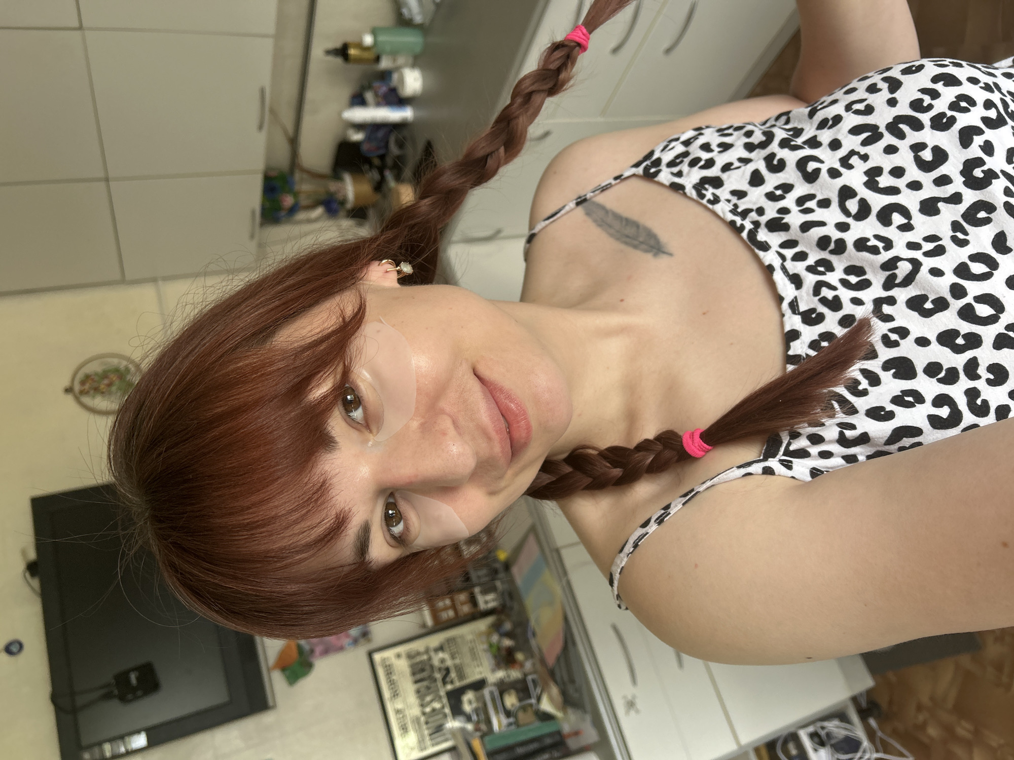 A woman with red hair and eye patches is looking at the camera. She has two braids tied with pink rubber bands. She is wearing a white tank top with black spots and has a tattoo of a feather on her left shoulder. The background shows a room with a TV, drawers, and various objects on a table. There is no information to identify the location as the city of Фоки, Russia.