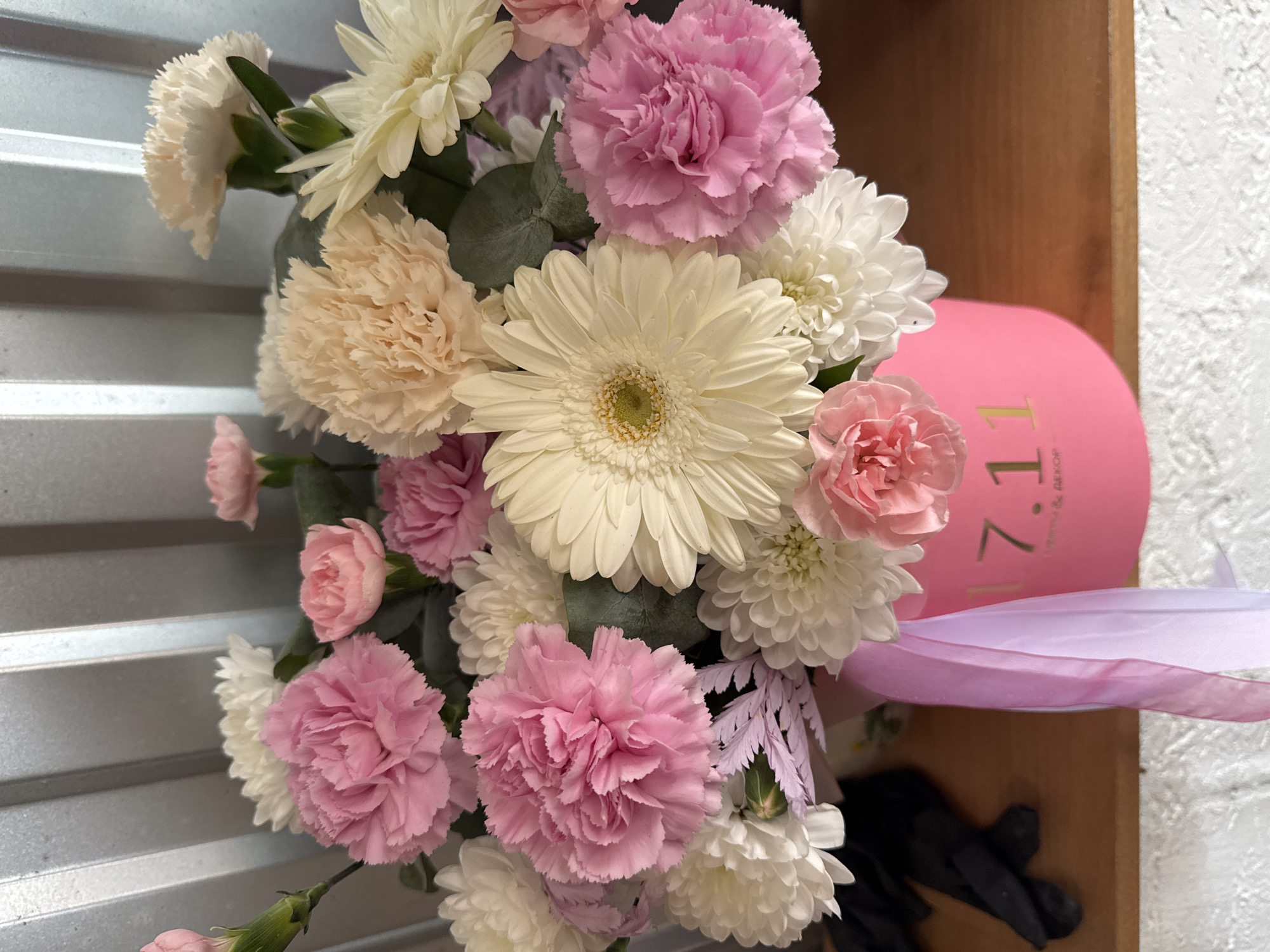 A floral arrangement in a pink cylindrical box is presented indoors. The arrangement includes white gerbera daisies, pink and white carnations, and white chrysanthemums. A light purple ribbon adorns the box. The setting appears to be near a wooden surface, possibly shelves. The image may have been captured near or in Ольховка, Russia, based on contextual clues.