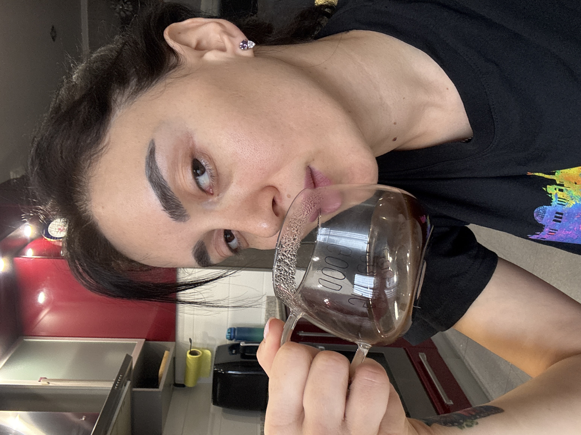 A woman is holding a glass mug with liquid in it in an indoor environment. The woman has dark hair, light skin, and is wearing a black t-shirt. The mug has the word "GOOD" written on it. The background shows a kitchen with red cabinets and stainless steel appliances. The exact location within Tbilisi, Georgia, cannot be determined from the image.