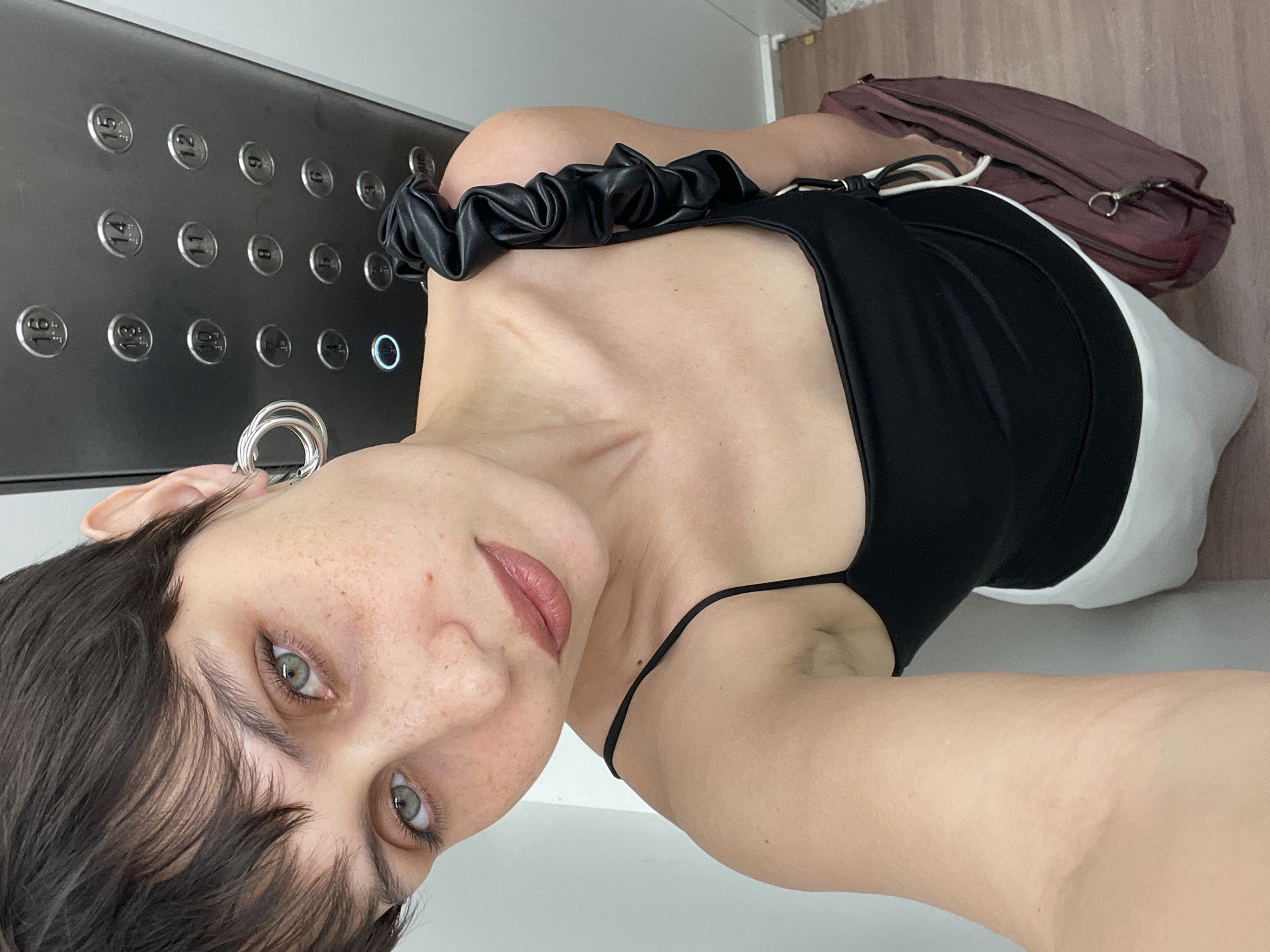 A woman with dark hair and fair skin is taking a selfie in an elevator. She is wearing a black spaghetti strap top and white shorts or a skirt. She has silver hoop earrings. Behind her are the elevator buttons, indicating floors 1 through 16 plus a zero floor. The elevator is illuminated with a blue light on the zero floor button. A maroon bag rests on the floor near the woman's right side. The floor of the elevator has a light brown wood-grain finish. The woman is the only person visible in the image, and there is no information to indicate the city of Izhevsk, Russia.