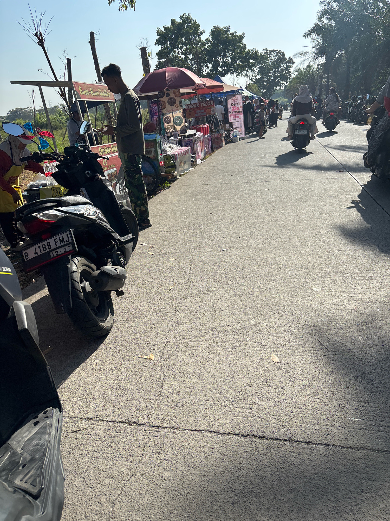 The image captures a busy street scene in Telagamurni, Indonesia, with vendors and customers lining the roadside. In the foreground, a black motorcycle with a license plate "B 4188 FMJ" is parked. To its left, a vendor in a yellow apron is attending to a customer, with various food items and drinks displayed on a small cart. Further down the street, a man in military-style camouflage pants is interacting with a vendor at a stall labeled "Bum-Bum Thai Tea." Several other stalls with umbrellas are set up along the road, selling goods like "Gerai Telor" (egg stall) and "Gerai Zebra Hamster." A line of motorcycles and people are moving along the street, indicating a lively marketplace. The environment is characterized by a clear blue sky and lush green trees in the background, suggesting a sunny day in a semi-urban or rural setting.