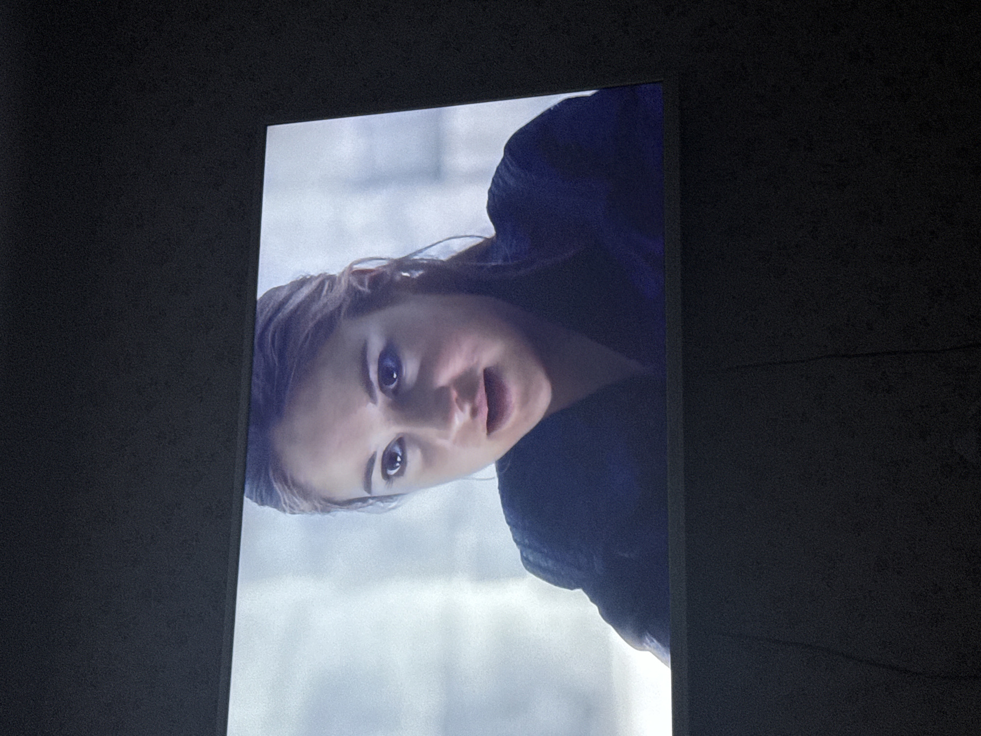 The image shows a close-up of a woman's face on a screen, likely a television or monitor, against a dark, neutral background. The woman has fair skin, light brown hair, and is wearing a dark jacket. She is looking directly at the viewer with a slightly open mouth, suggesting she might be speaking or expressing surprise. The background of the image displayed on the screen appears to be a neutral, possibly urban, setting with indistinct grey and white tones. The image was likely taken indoors in a place to watch TV. There is no evidence to suggest this image was taken in Mytishchi, Russia.