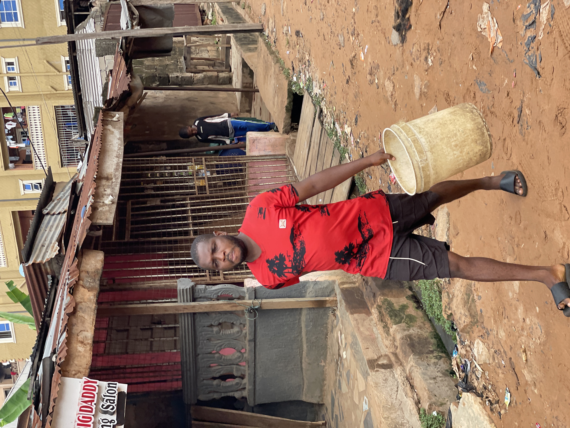 A man stands on a muddy, litter-strewn ground in Onitsha, Nigeria, holding an empty, off-white plastic bucket. He is wearing a red T-shirt with a black tree-like print, black shorts, and black sandals. He is facing the camera. In the background, there are buildings constructed with corrugated iron and concrete. A building on the left has a sign indicating "BIG DADDY og Salon." Another person is visible further back under a rudimentary shelter. The surrounding environment shows signs of decay and poverty.