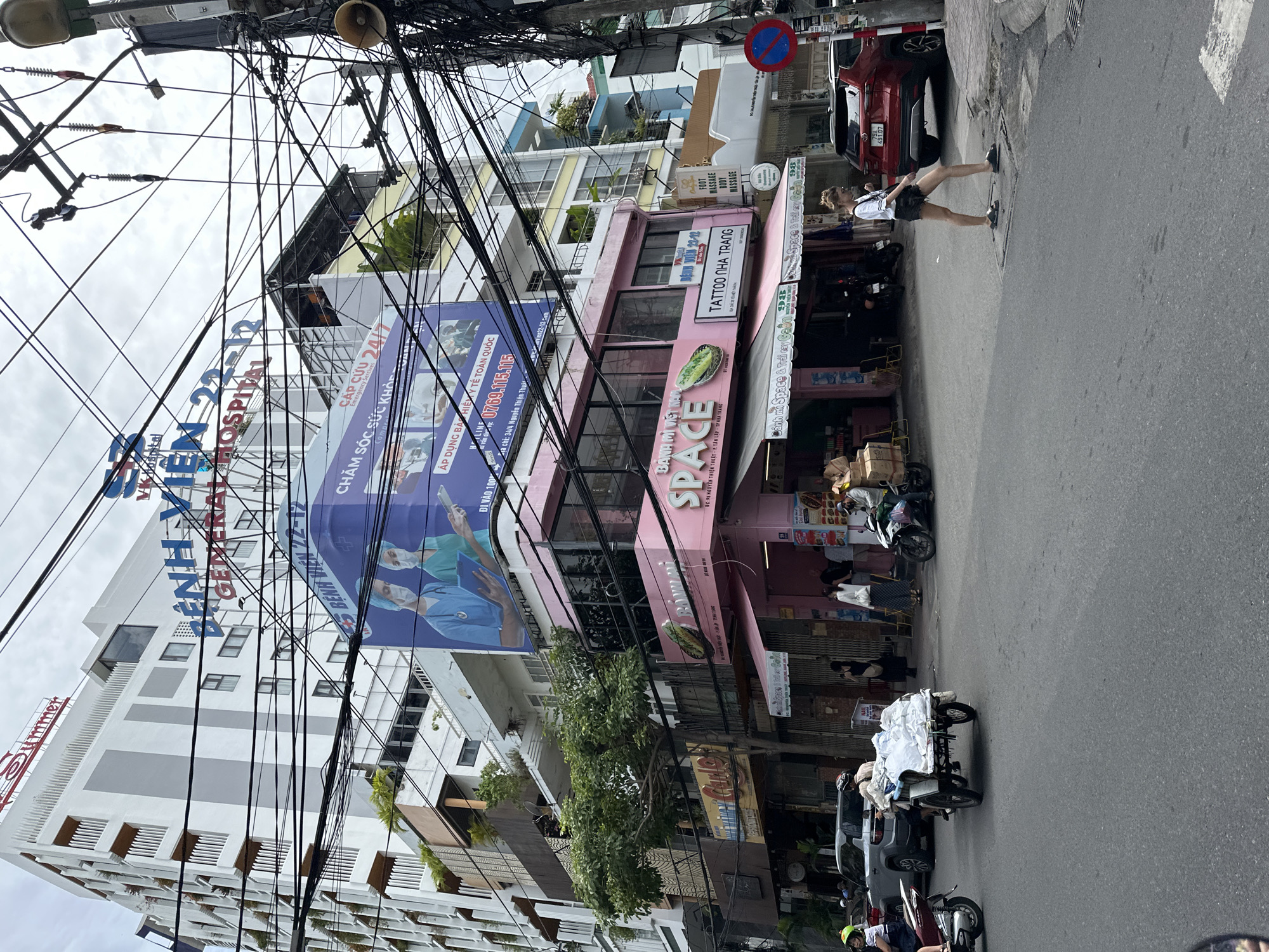 In Nha Trang, Vietnam, a street scene unfolds under an overcast sky, dominated by a complex web of overhead electrical wires. A tall white building, labeled "BỆNH VIỆN 22-12 GENERAL HOSPITAL," stands prominently in the background, its facade adorned with vertical grey stripes and numerous windows. Below the hospital, a large blue billboard features an advertisement for "CẤP CỨU 24/7" (Emergency 24/7) with images of medical professionals.
The foreground is occupied by a pink building with "SPACE" and "BÁNH MÌ VIỆT NAM" signage. Adjacent to this, a shop called "TATTOO NHA TRANG" is visible. Several people are present: a woman in black shorts and a white t-shirt walks on the sidewalk towards a red SUV parked on the street. A cyclist on a motorcycle with a cart full of white goods moves past. Another motorcycle is parked in front of the pink building. Various other pedestrians and parked vehicles, including a silver SUV and multiple motorcycles, are scattered throughout the scene. The environment is a typical urban street with paved roads and sidewalks, interspersed with greenery from trees and planters. A red and blue circular traffic sign is affixed to a pole on the right side of the image.