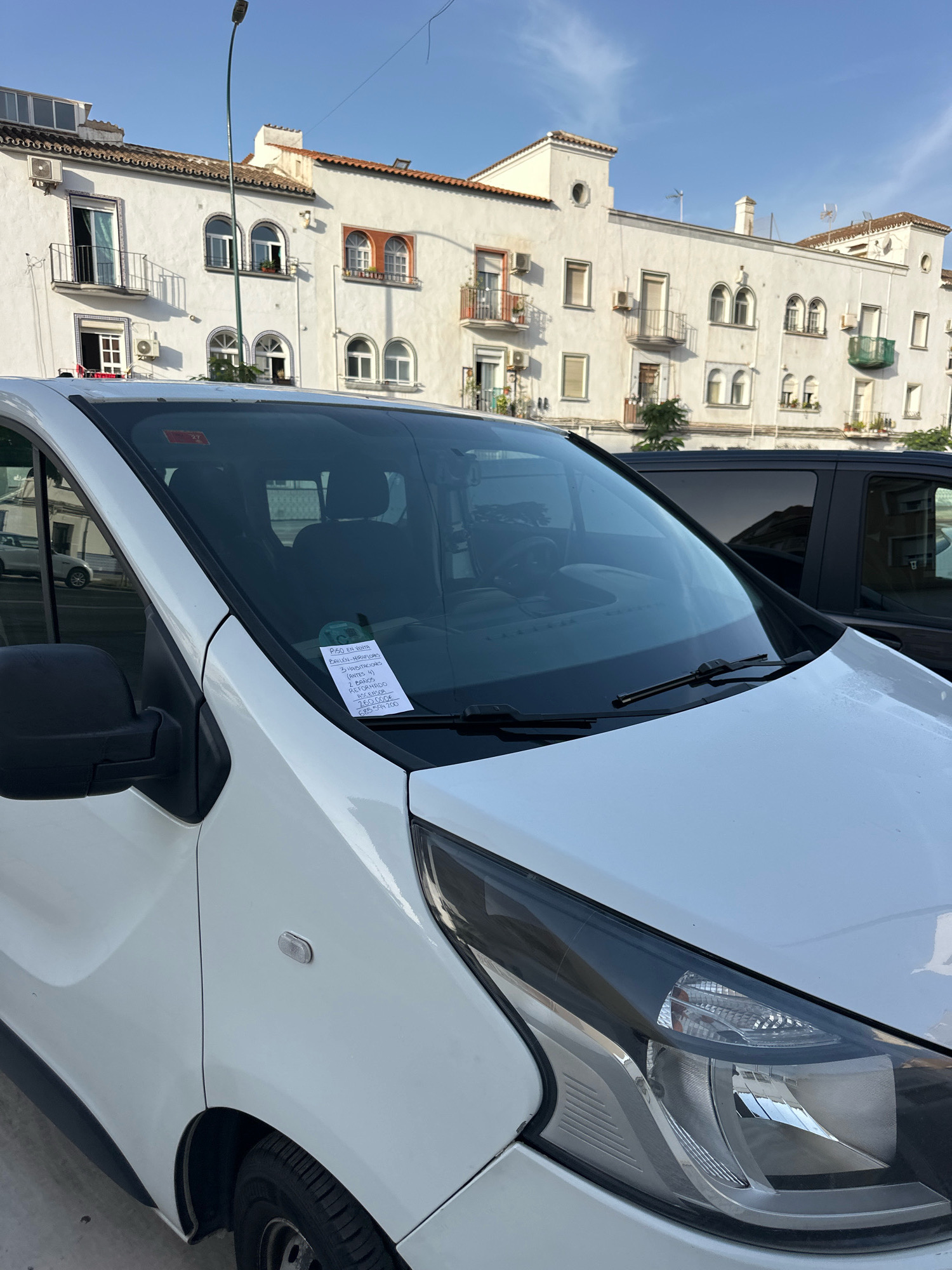 The image shows the front passenger side of a white van parked on a street in Malaga, Spain. On the windshield, there is a parking ticket or notice, and a red sticker is visible in the upper corner. The van is parked in front of a white building with multiple windows and balconies. The sky is blue with some clouds. In the background, another dark-colored vehicle is partially visible. The street appears to be paved.