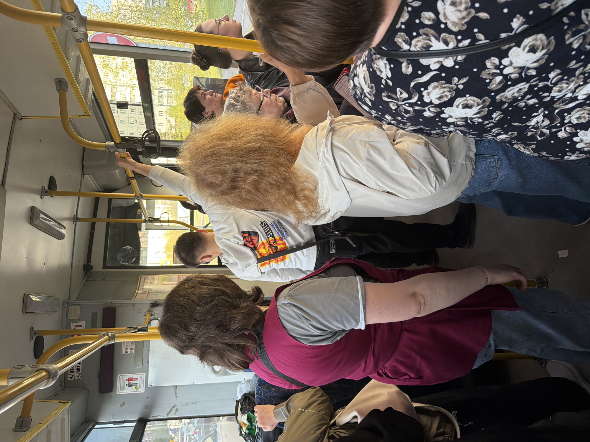A bus in Krasnoyarsk, Russia, is carrying multiple passengers. The interior features yellow handrails along the aisle and near the windows. The passengers are standing and sitting, varying in age and attire. A woman in the foreground wears a maroon vest over a gray top, while another individual is wearing a white jacket and blue jeans. The environment suggests an urban setting with buildings visible through the bus windows.