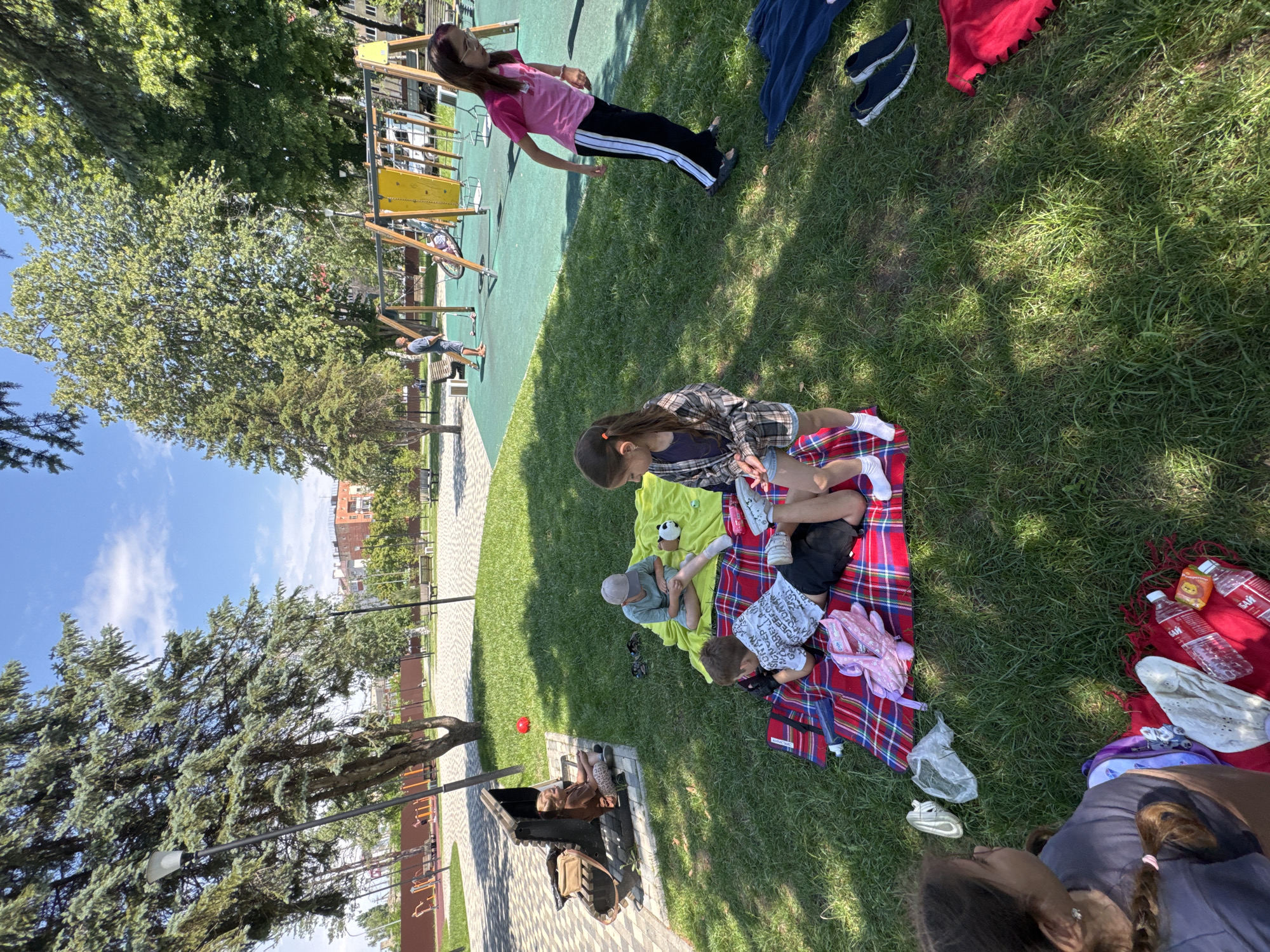 A group of people are having a picnic on a grassy area near a playground. The scene is outdoors on a sunny day. The subjects include several children and adults. One child, a girl, is wearing a pink shirt and black pants with white stripes, and she is standing. Another child, also a girl, is sitting on a red and blue plaid blanket, wearing a plaid shirt and denim shorts. A young boy is sitting on a yellow blanket, wearing a light blue shirt and grey cap. A man is sitting on the ground near a playground structure. Another child is lying on the plaid blanket, wearing a white shirt with text. Several water bottles are visible on a red blanket, with one bottle displaying text that appears to be in Russian. One of the water bottles has a yellow label with an image of a character. There is also a red ball on the grass. The location appears to be a park or recreational area, with trees and a playground visible in the background. The time of day seems to be midday, given the bright sunlight and shadows. The overall mood seems relaxed and leisurely.