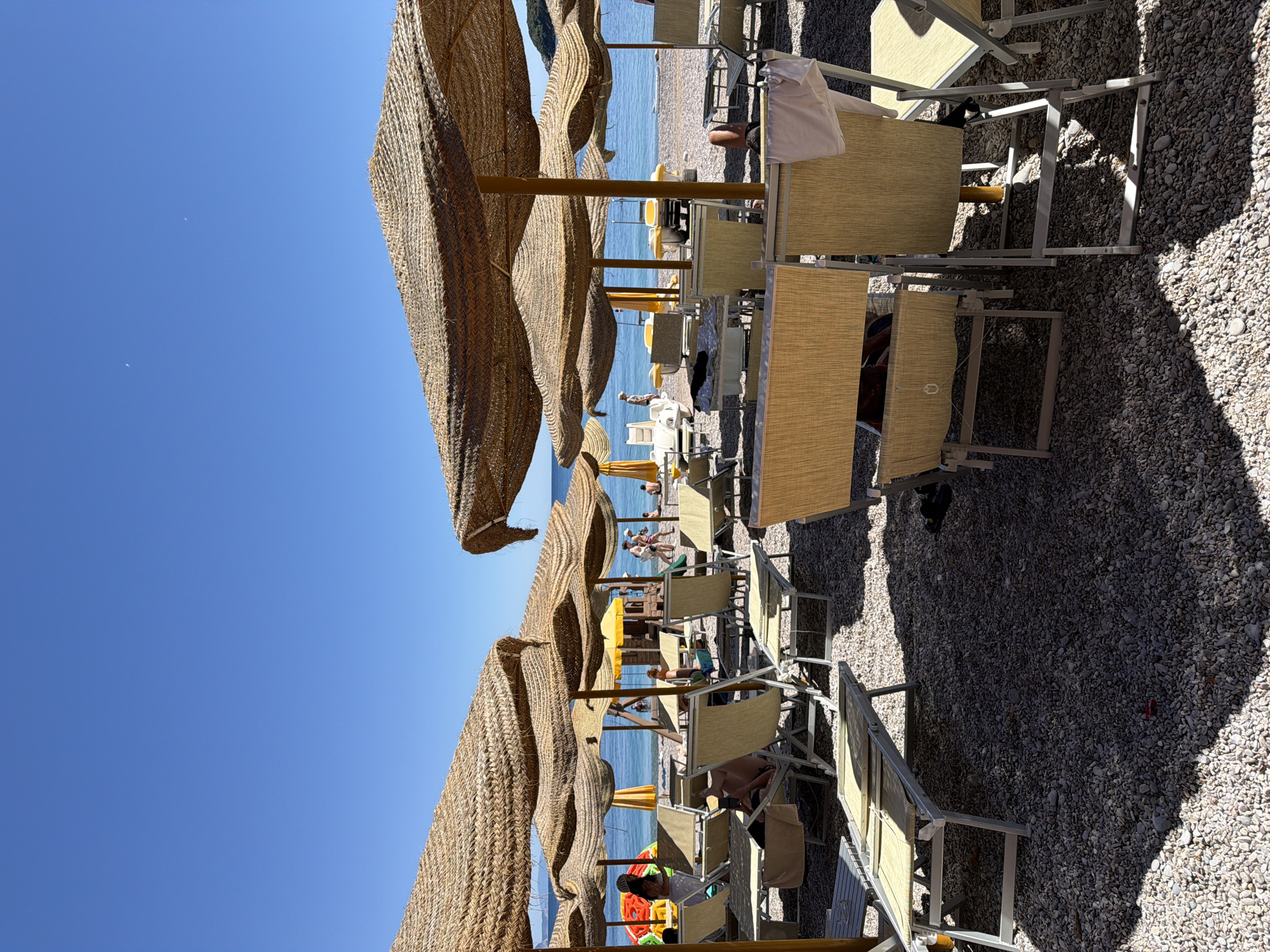 The scene depicts a beach lined with numerous sun loungers and straw parasols. The loungers, made of light-colored canvas stretched over metal frames, are mostly unoccupied, casting shadows on the pebbled beach. Patches of sunlight break through the shade of the parasols. The beach slopes gently towards the blue Adriatic Sea, with a line of people and structures visible in the distance. The sky above is clear and a vivid blue. The orientation of the photograph suggests it was taken vertically. Based on the provided location, the scene is located at a beach at or near the city of Prijevor, Montenegro.