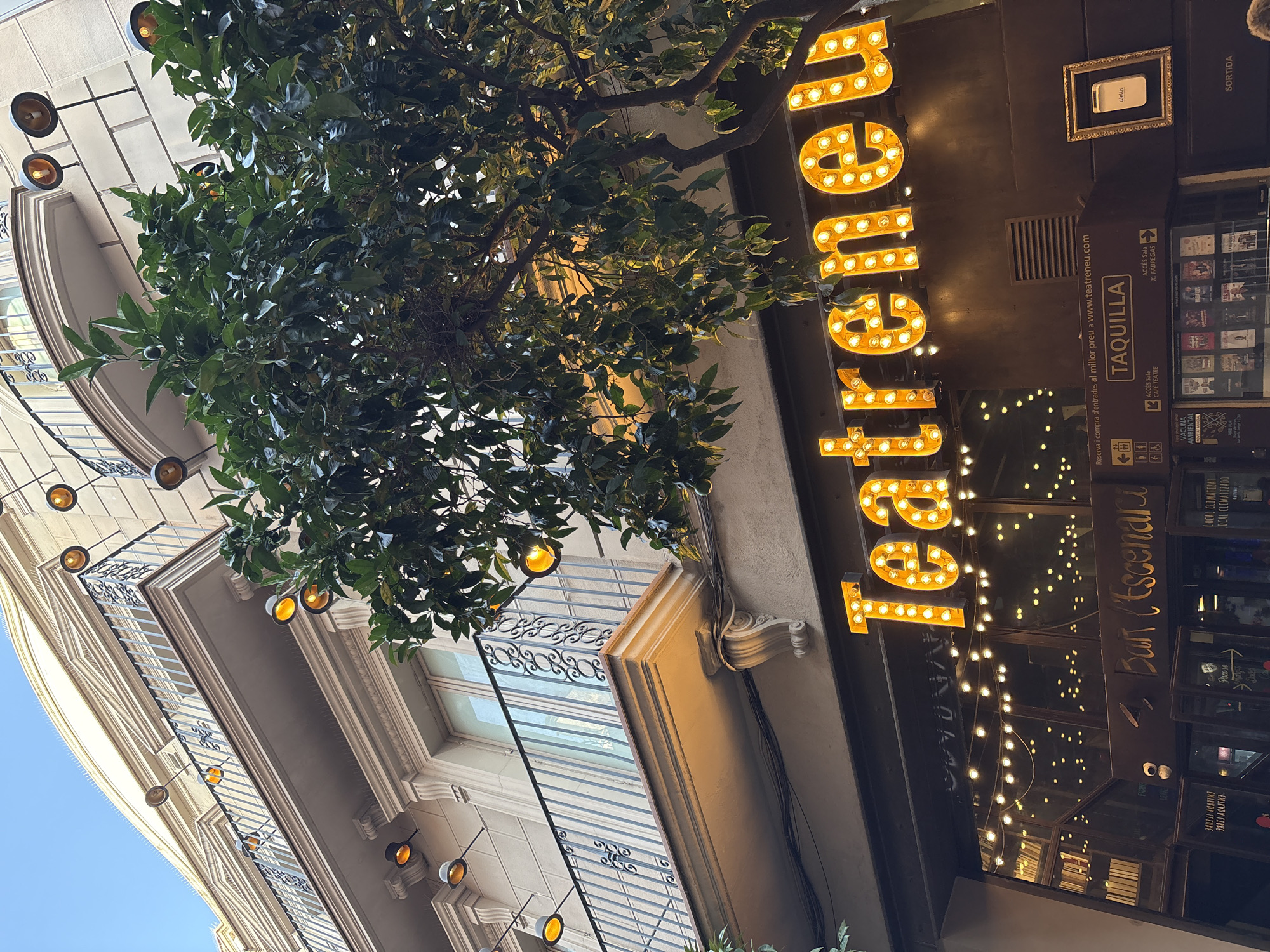 The image shows the facade of a building in Barcelona, Spain, featuring a theater called "Teatrenou." The building has a light-colored stone facade with balconies and ornate railings. A tree with green leaves and some small green fruits is positioned in front of the building, partially obscuring the upper floors. String lights with exposed bulbs hang across the front of the building, illuminating the signage. The main sign for "Teatrenou" is made of large, illuminated letters. Below this, there is a ticketing booth area with additional signage and posters displayed in a glass-fronted display case. The entrance to the theater and bar is visible, with open doors leading inside. The overall impression is of a vibrant cultural venue in an urban setting.
