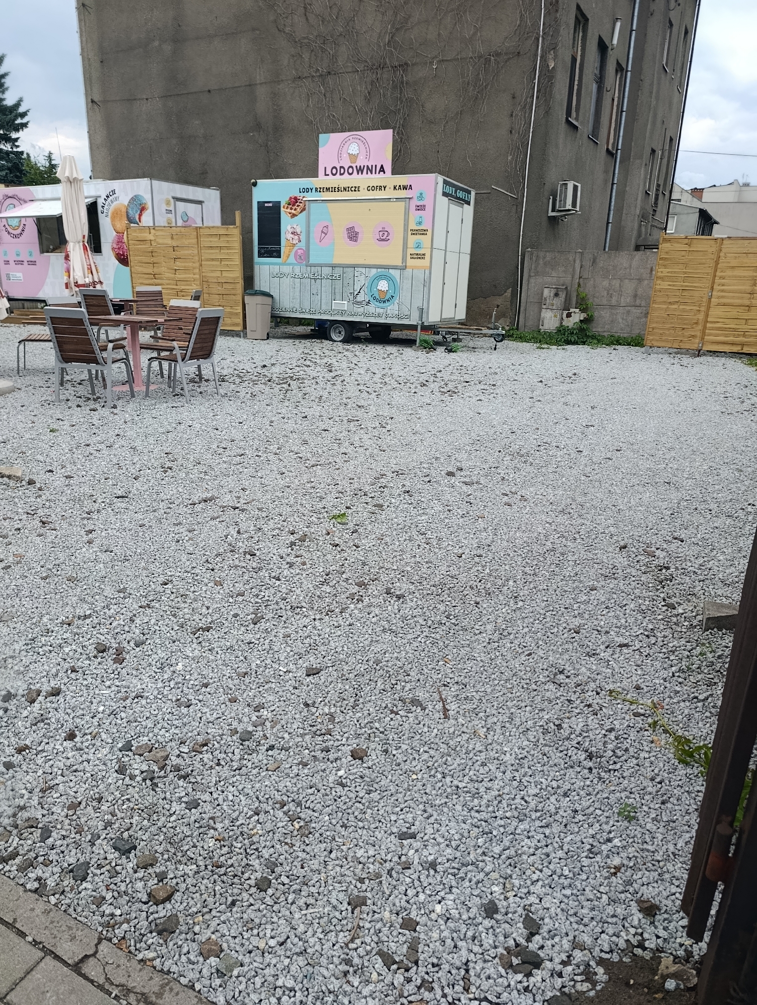 The image shows an outdoor scene in a city. In the foreground, a gravel-covered area dominates the view, with some scattered larger stones and a concrete border on the left. In the mid-ground, a seating area with a table and chairs is visible, positioned on the gravel. To the right of the seating area, there are two food trailers, one pink and one blue, with colorful graphics and text. The pink trailer has text that reads "PONCZKOWA" and features images of donuts. The blue trailer is labeled "LODOWNIA" and displays images of ice cream, waffles, and coffee, with text indicating "LODY RZEMIEŚLNICZE" (craft ice cream) and "GOFRY - KAWA" (waffles - coffee). Behind the food trailers, a tall, weathered grey building with visible ivy growth and boarded-up windows stands prominently. A wooden fence is also present to the right of the trailers. The environment appears to be an urban outdoor space, possibly a plaza or a lot next to a building, with a focus on the food vending operation.