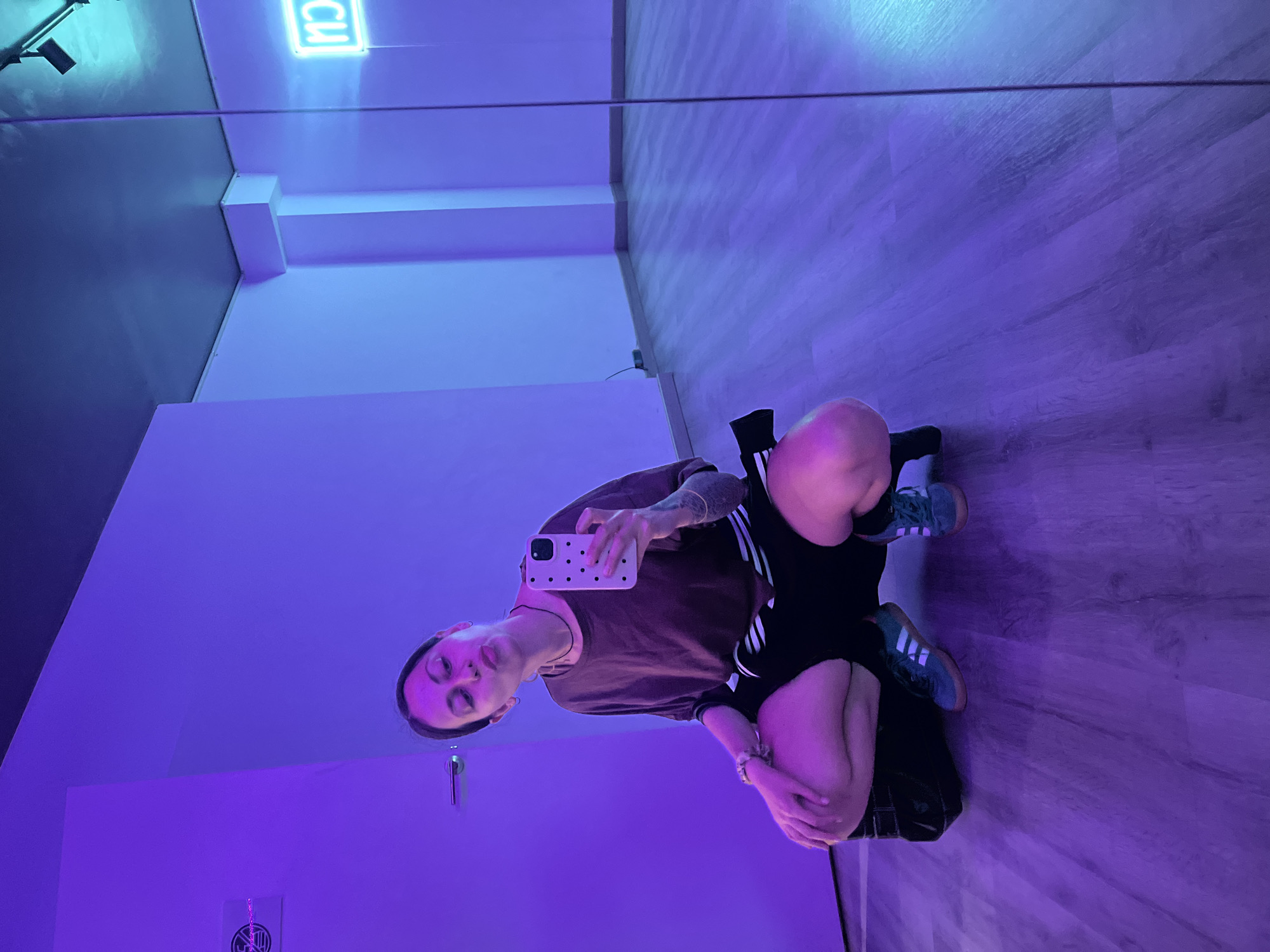 This media file is an indoor mirror selfie, likely taken in a studio or practice room, set in Barcelona, Spain. The scene is illuminated by striking purple and blue neon lighting, creating a moody and atmospheric ambiance.
The primary subject is a person, appearing to be female, reflected in a large mirror. She is in a squatting or low sitting pose with her knees bent, holding a white smartphone with a black polka-dot case to capture the image. She is dressed in a plain brown short-sleeved t-shirt, black athletic shorts with white stripes (reminiscent of Adidas design), light-colored socks, and dark sneakers also featuring white stripes. Notable details include tattoos on her right arm and shoulder, multiple rings on her fingers, and a pensive or neutral expression on her face. Her dark hair is pulled back.
The room's environment, visible in the reflection, features a wooden laminate floor and light-colored walls. In the upper left corner of the reflection, a bright cyan or blue light source is visible. Above the subject's reflected head, a neon sign displays the reversed letters "CU," indicating it might be part of a larger word or name. Below this, near the bottom of a white panel, there's a small, dark, circular logo or emblem, though its specific details are indistinct. The predominant artificial lighting suggests the picture was taken during the evening or night. The activity depicted is a casual self-portrait taken in a reflective surface.