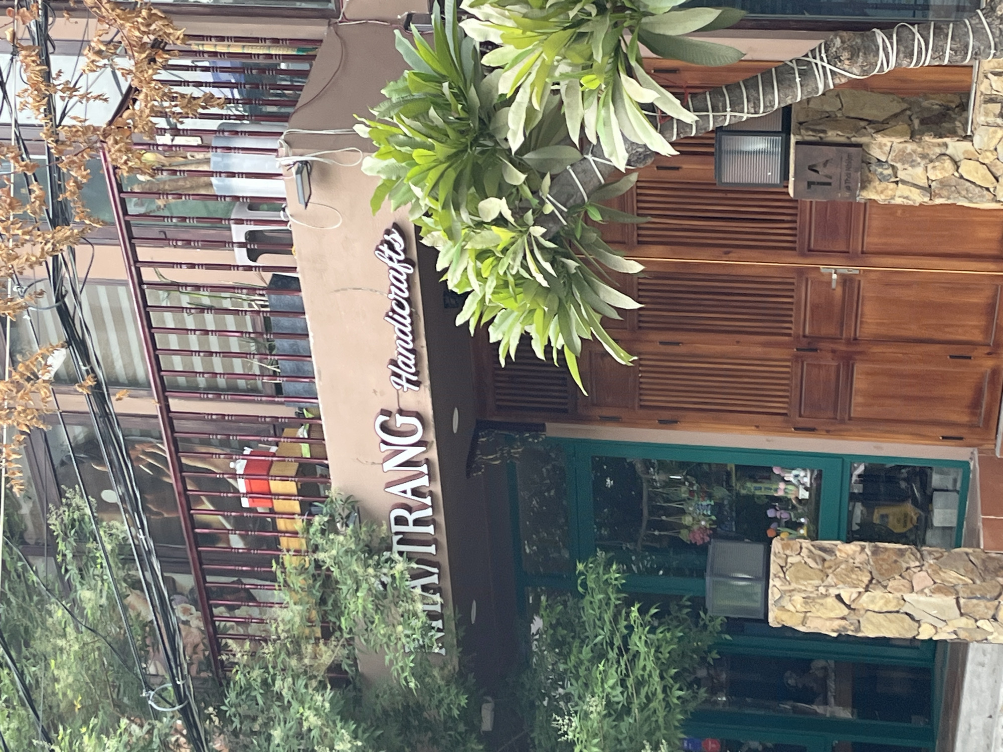 The image shows a storefront in Nha Trang, Vietnam, with the name "NHA TRANG Handicrafts" illuminated in white lettering on a tan facade. The storefront has a large green-framed window displaying various items, and a wooden double door. A stone pillar stands in front of the entrance, topped with a black light fixture. Lush green foliage, including a large tree with white-wrapped branches, partially obscures the view. A balcony with a brown railing is visible on the upper level of the building. The overall impression is that of a shop selling local crafts.